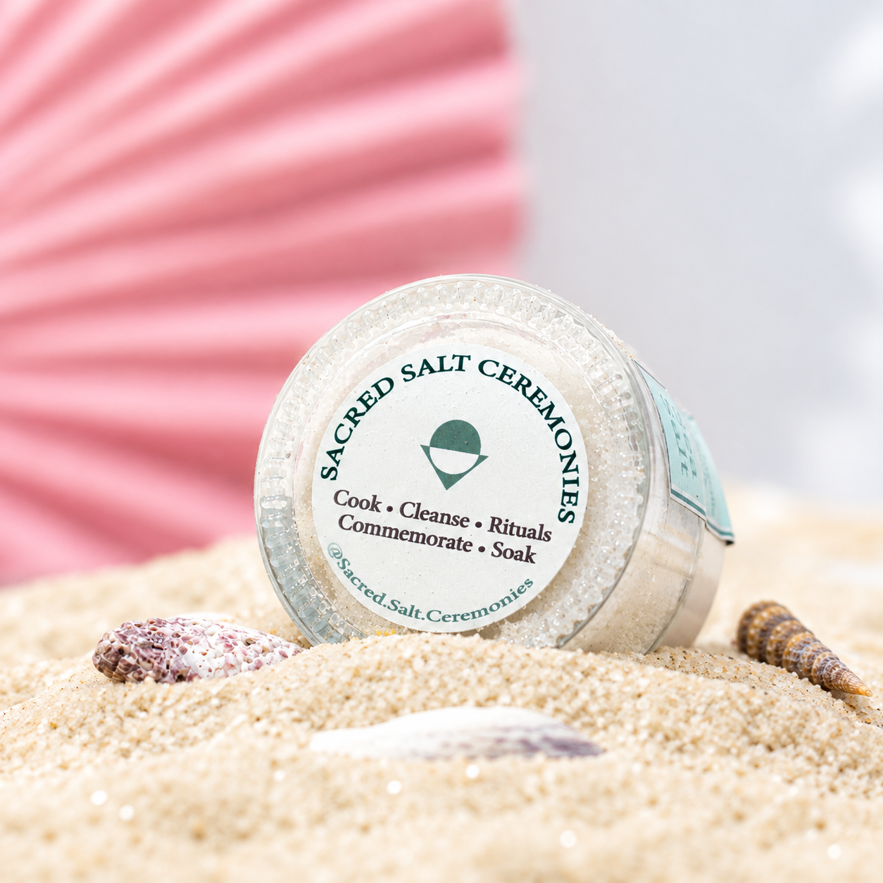 A container of Sacred Salt Ceremonies salt surrounded by sand and seashells, with a pink paper fan in the background.