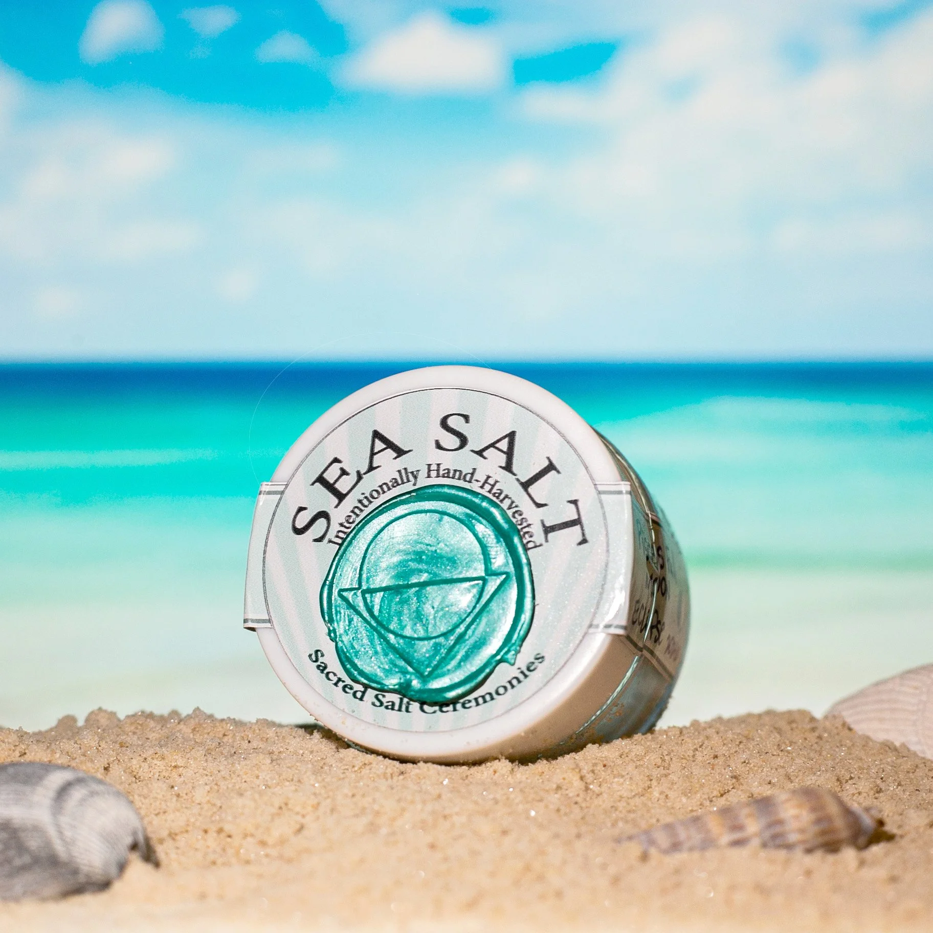 A small container of sacred salt with the label 'Sacred Salt, Naturally Hand-Harvested, Sacred Salt Ceremonies,' sitting on sandy ground with shells.