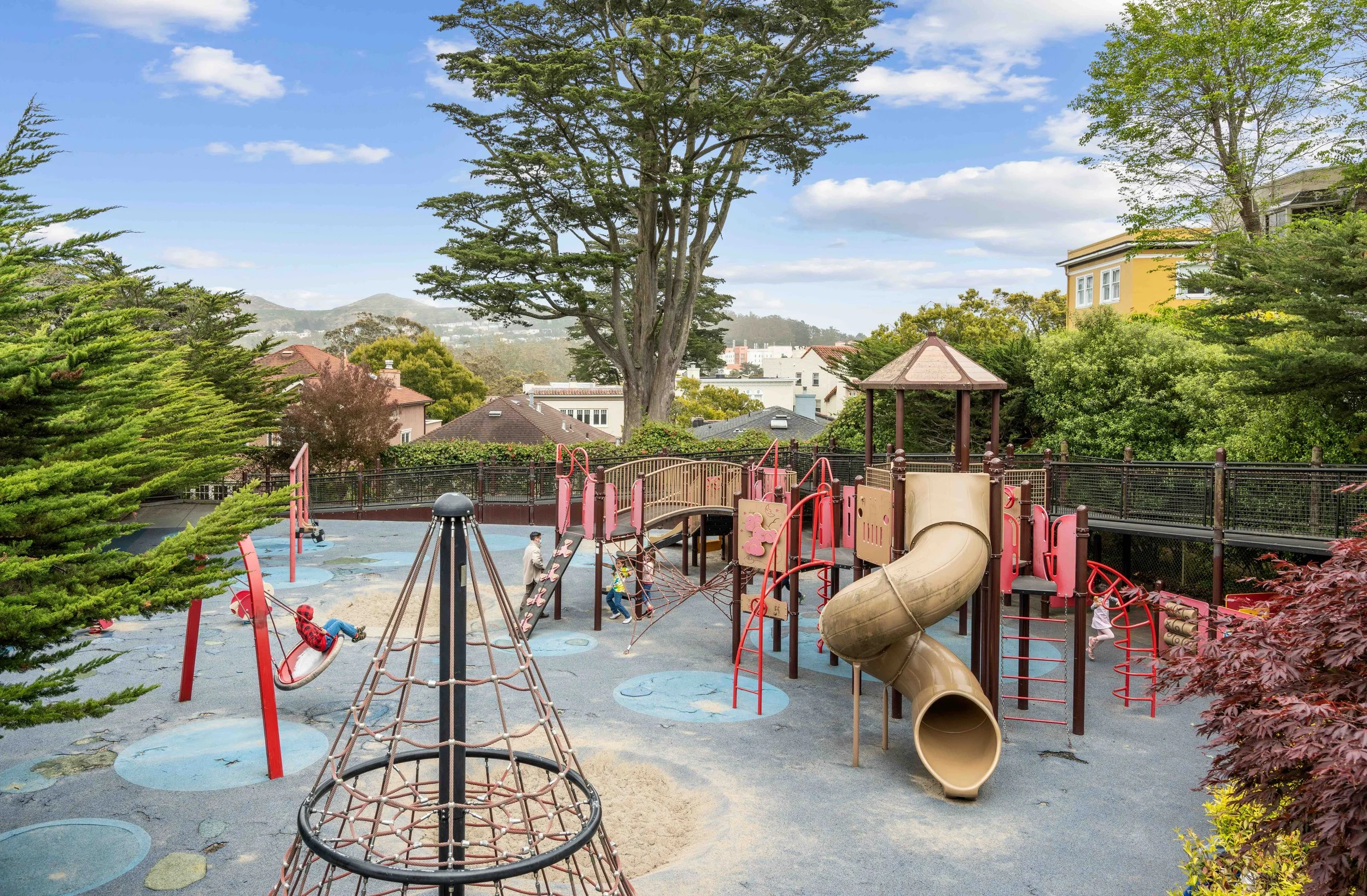 Children playing on a colorful playground with a slide, climbing structures, and swings, surrounded by trees and residential houses under a partly cloudy sky.