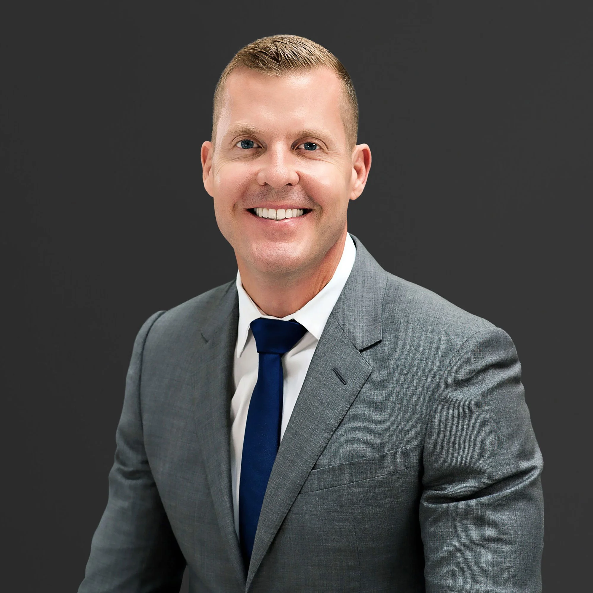 Professional portrait of a smiling man in a gray suit, white shirt, and navy tie, against a dark gray background.
