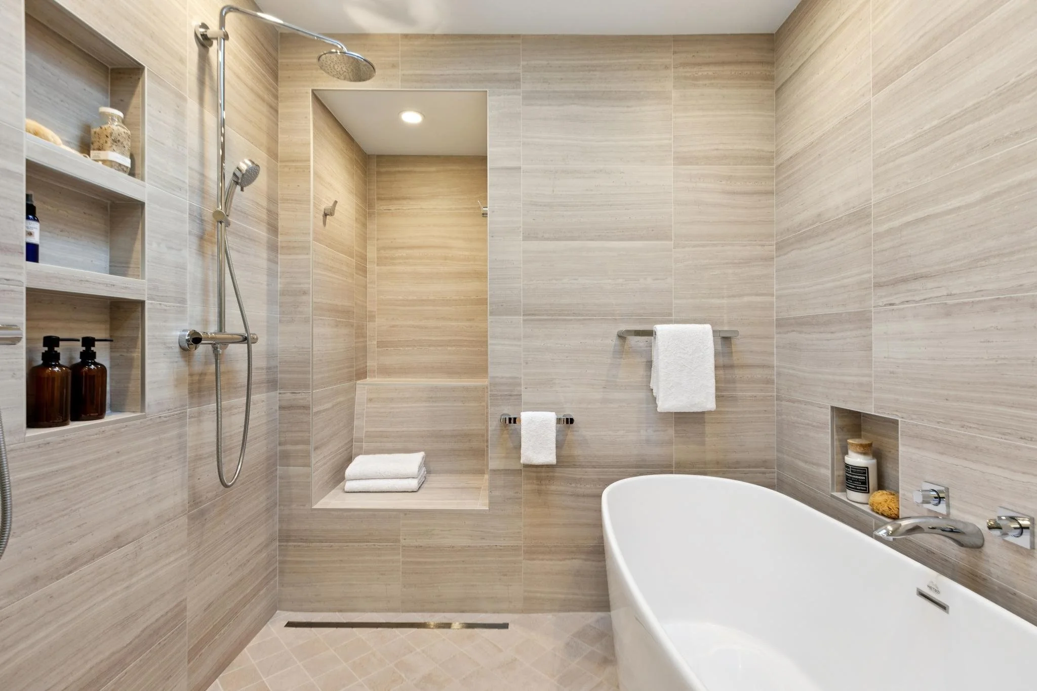 Modern bathroom with beige tiled walls and a white bathtub, featuring shelves with toiletries and a shower area with a rain shower head.