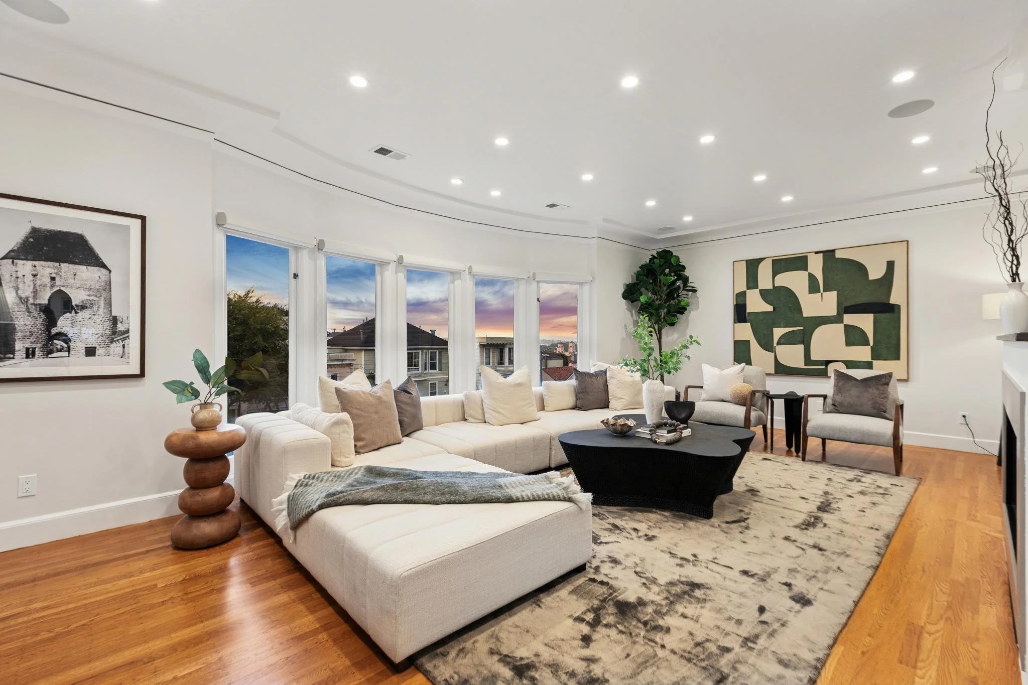 Bright, modern living room with a large white sectional sofa, multiple cushions, a black coffee table, and two armchairs. Floor-to-ceiling windows with white curtains show a sunset view. Decor includes a black-and-white framed photo, green potted pla