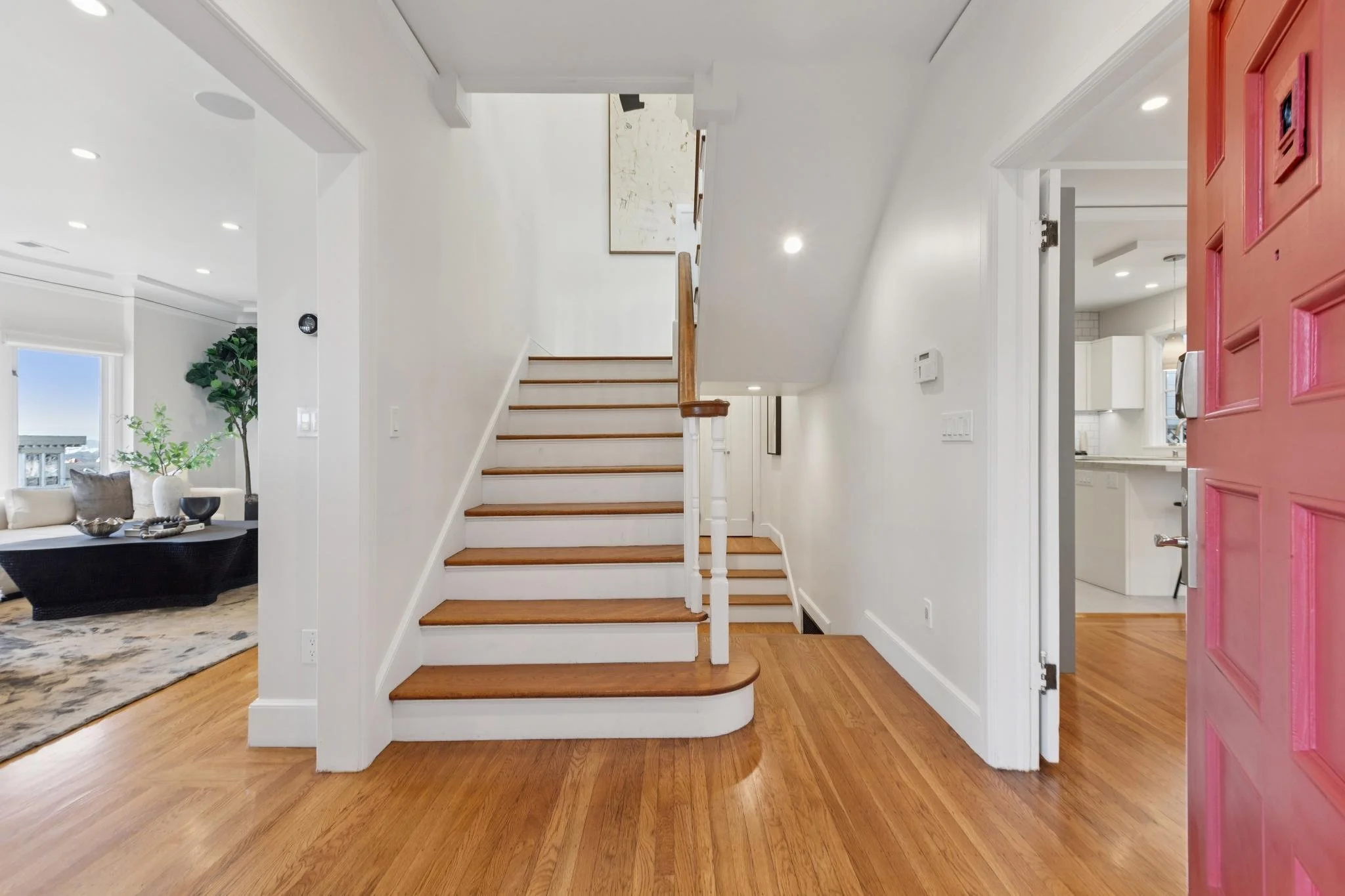 Interior view of a house entryway with wooden stairs, hardwood floor, white walls, a glimpse of the living room on the left, and the kitchen partially visible on the right, with a pink front door.
