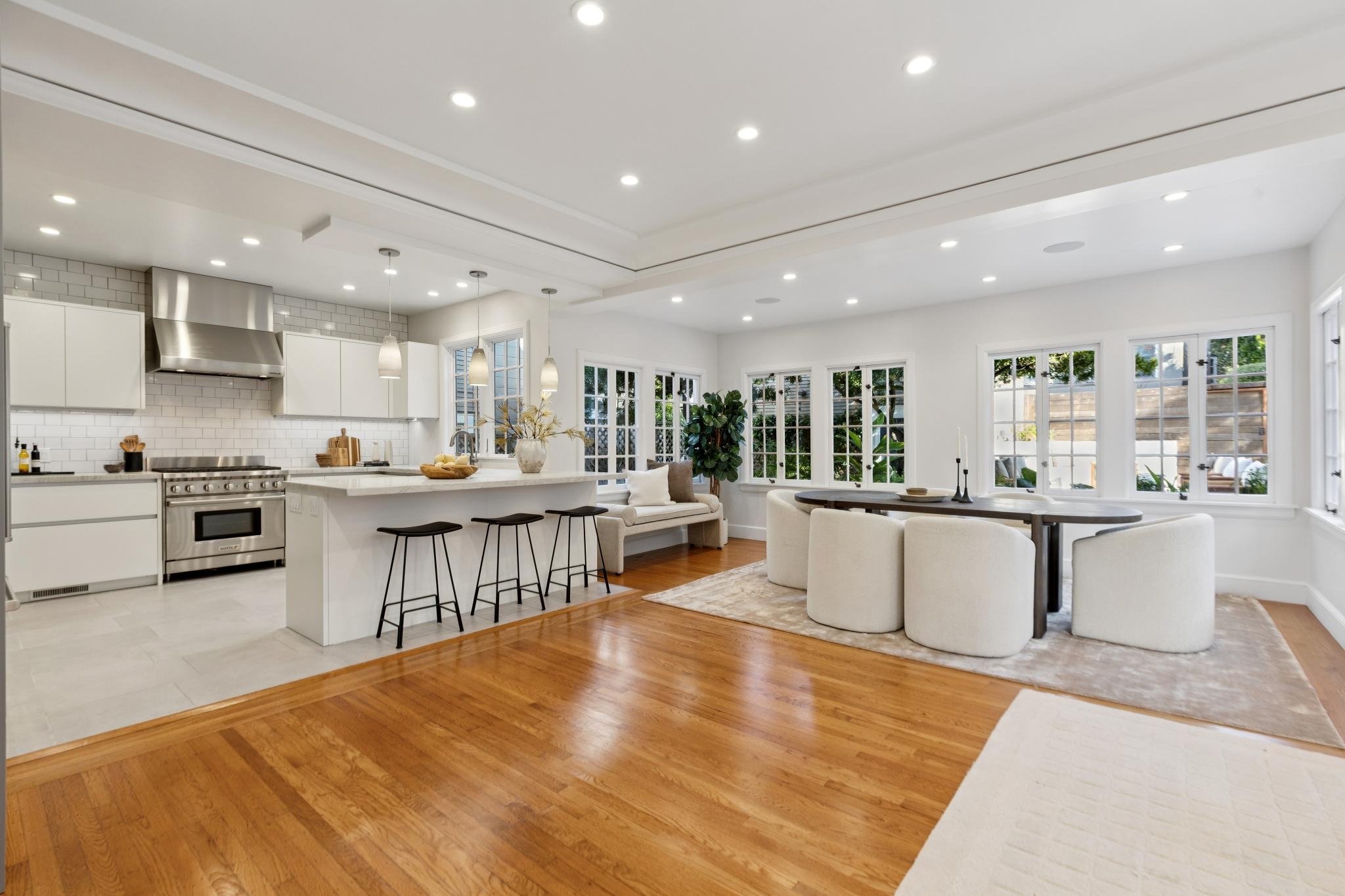 Modern open-concept kitchen and dining area with white cabinetry, stainless steel appliances, a white island with three bar stools, and a dining table with cream-colored chairs in a well-lit room with large windows.