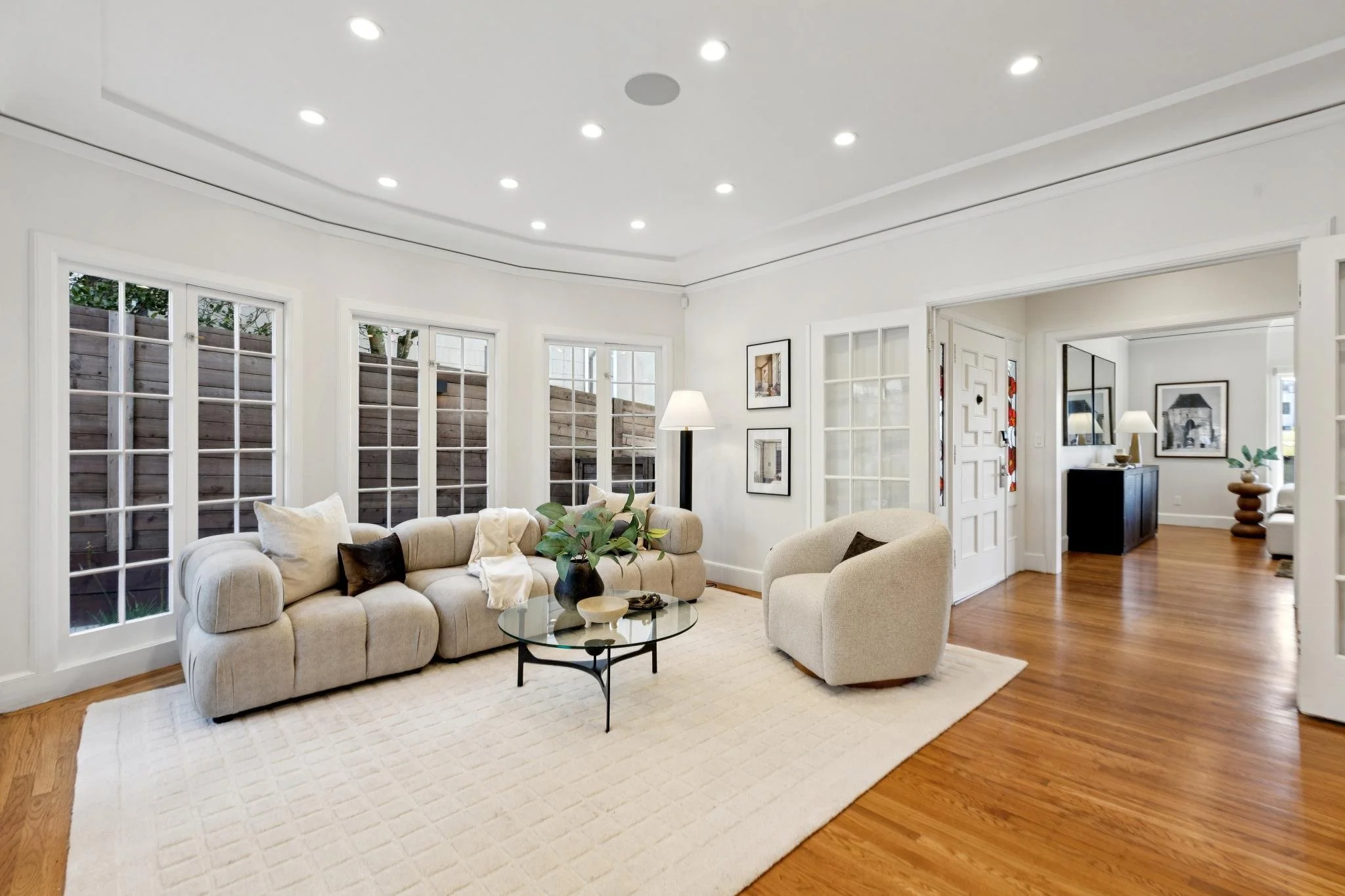 Living room with large windows, beige sofa, armchair, glass coffee table, tree in vase, white walls, hardwood floor, and framed artwork.