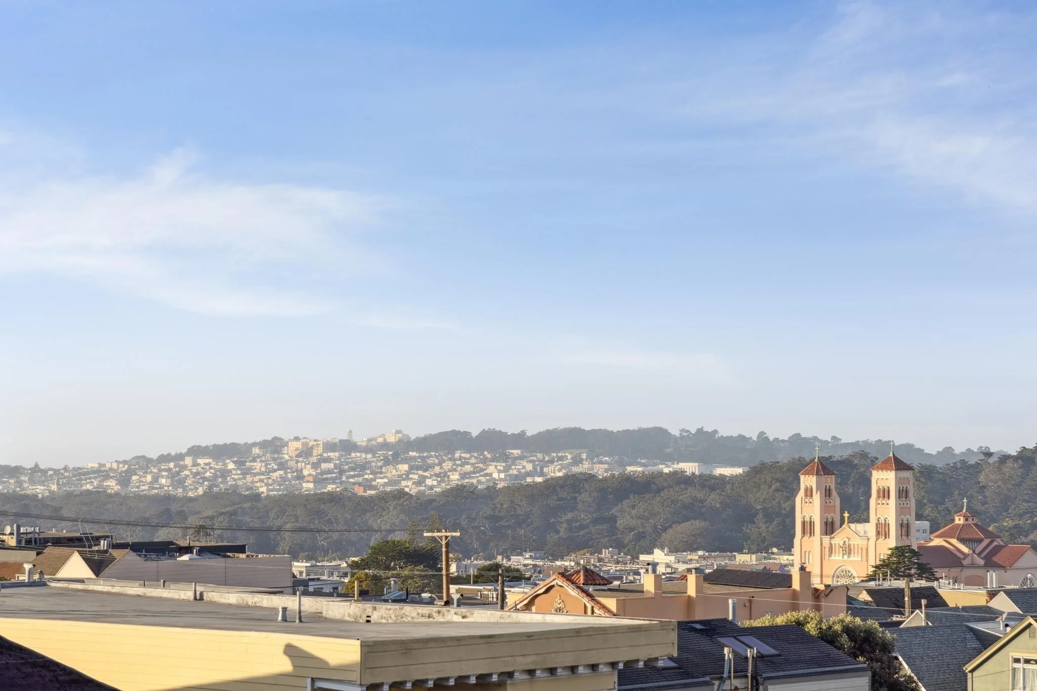 Cityscape with residential buildings, a church with twin towers, and a hill in the distance under a partly cloudy sky.