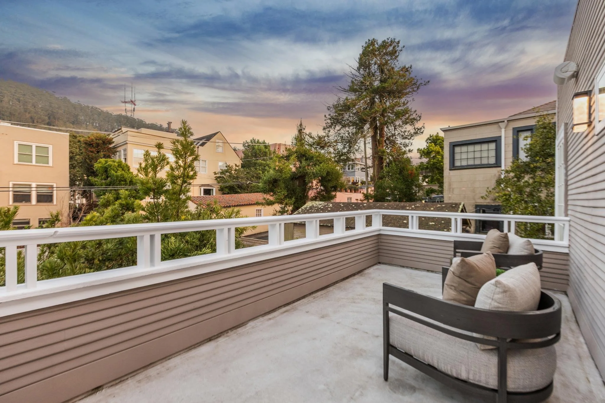 View of a balcony with outdoor seating, overlooking neighboring houses and trees during sunset.