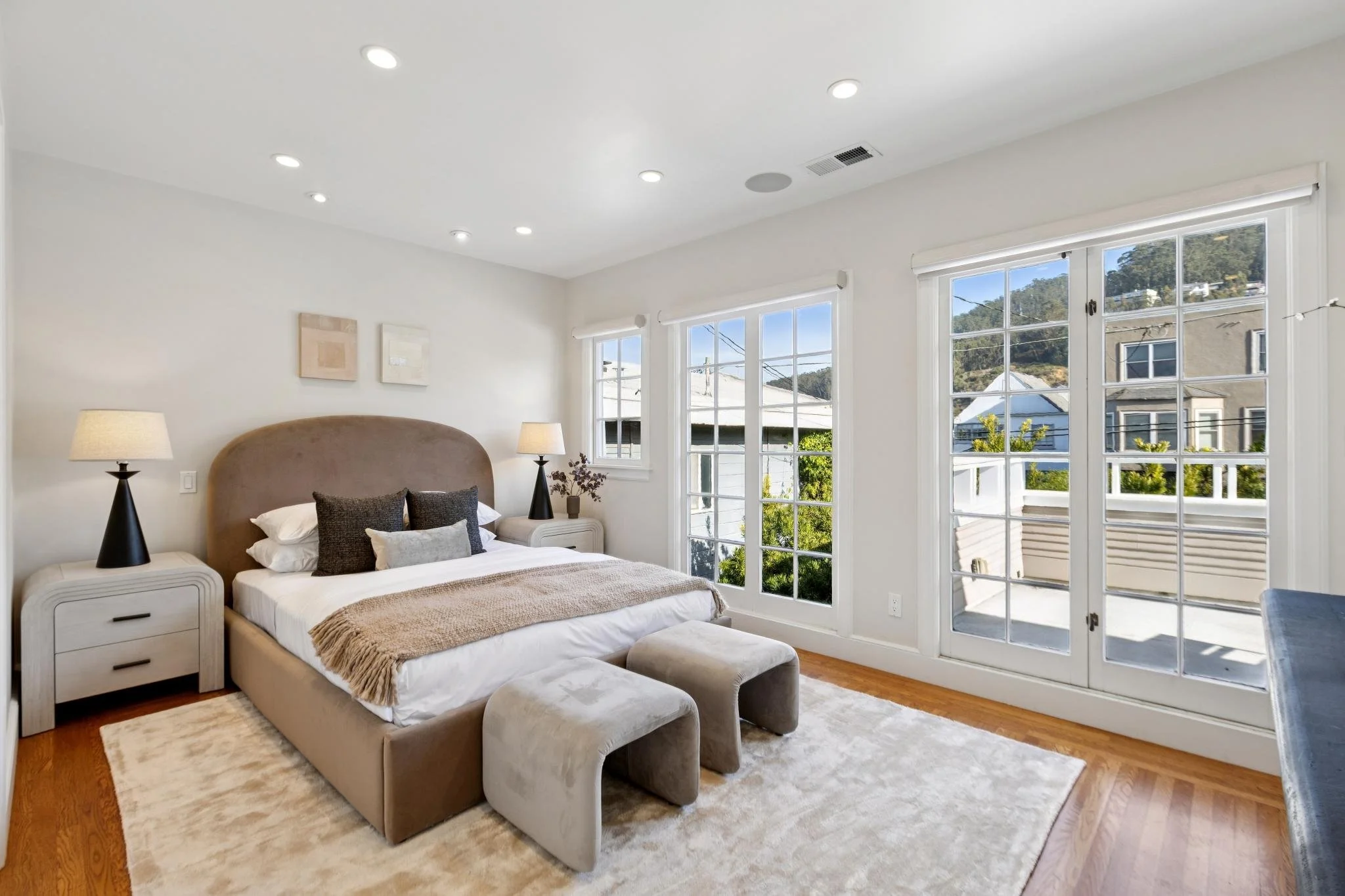 Bright bedroom with beige upholstered bed, two nightstands with lamps, and large windows showing a view of neighboring houses and trees.