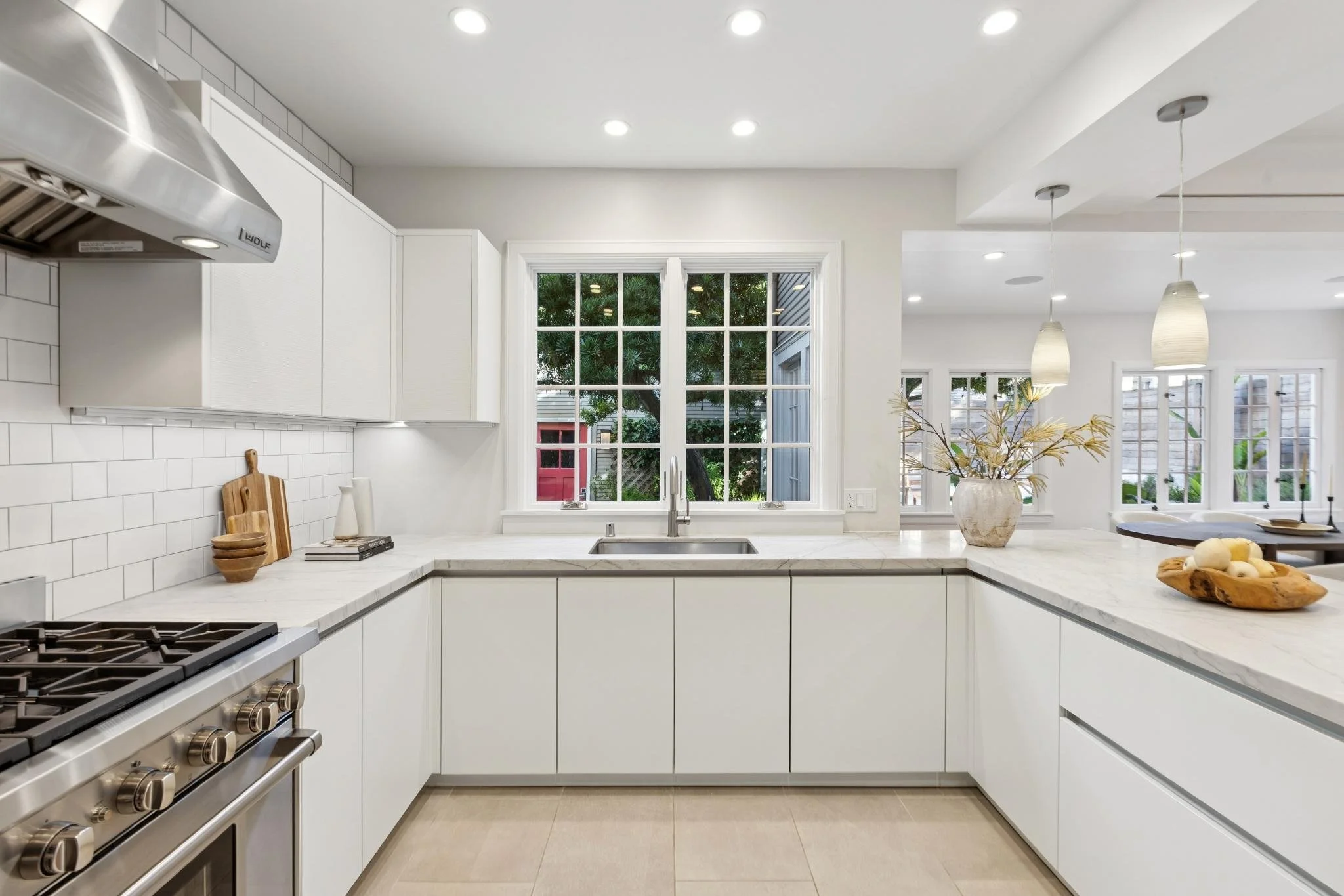 White kitchen with large window, marble countertops, and a stainless steel stove, decorated with wooden bowls and a vase of flowers.