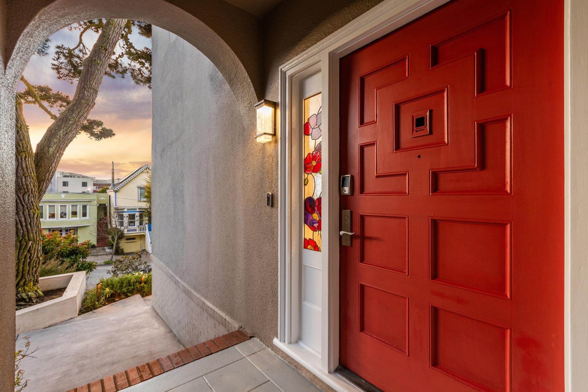 Red front door with decorative geometric panels, side stained glass window with floral design, and a wall-mounted light fixture at dusk, overlooking neighborhood houses and a large tree.