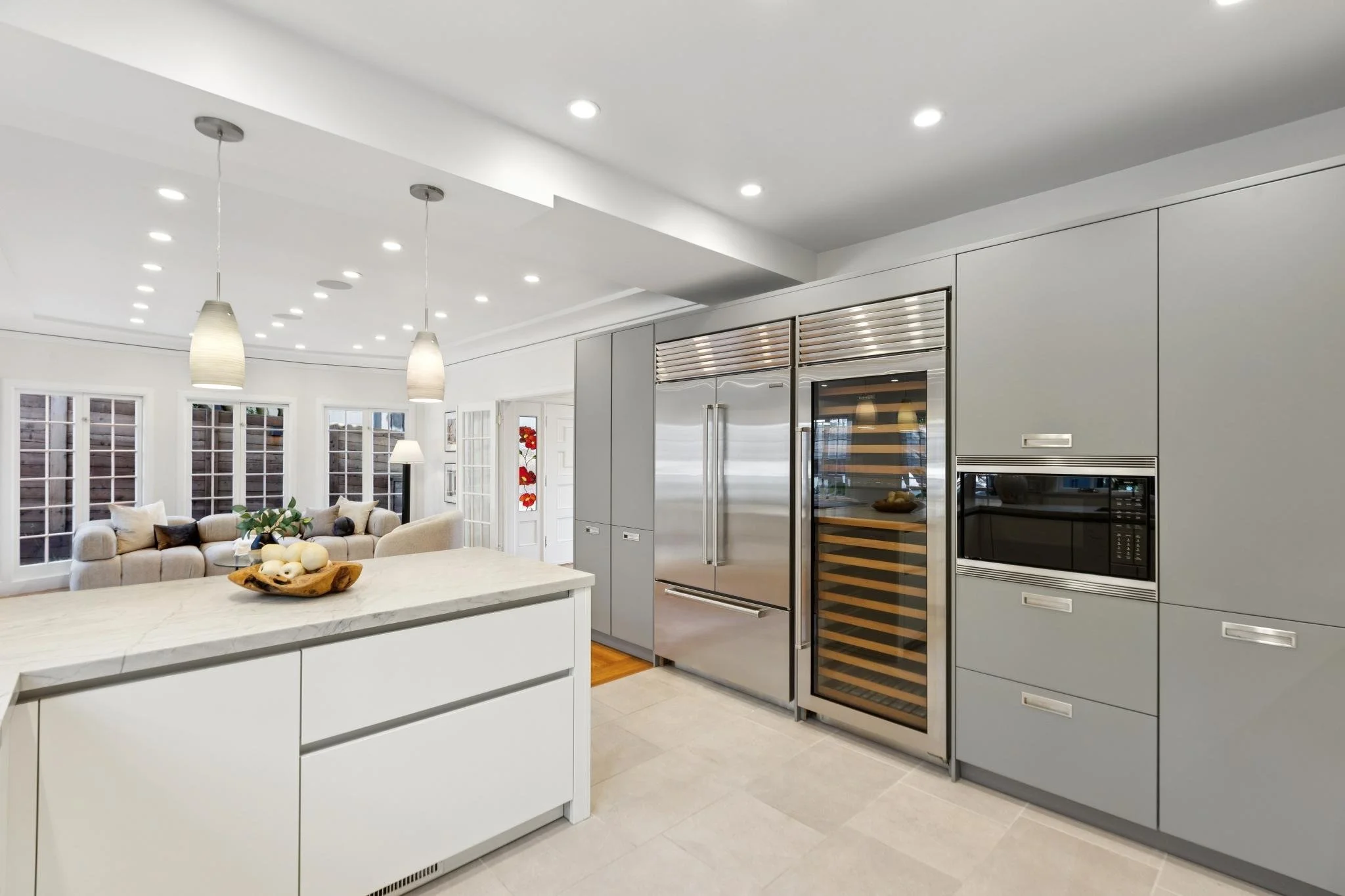 Modern kitchen with gray cabinetry, stainless steel refrigerator, wine cooler, microwave, white island with decor, and open living room with sectional sofa and windows