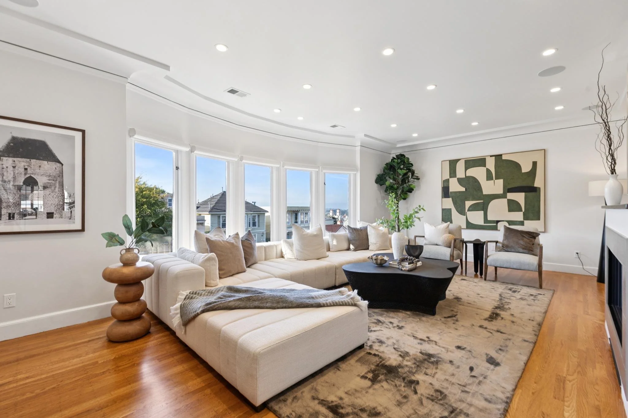 Bright living room with large sectional sofa, wooden side table with plant, abstract art on wall, potted plant, and coffee table, overlooking neighborhood houses through windows.