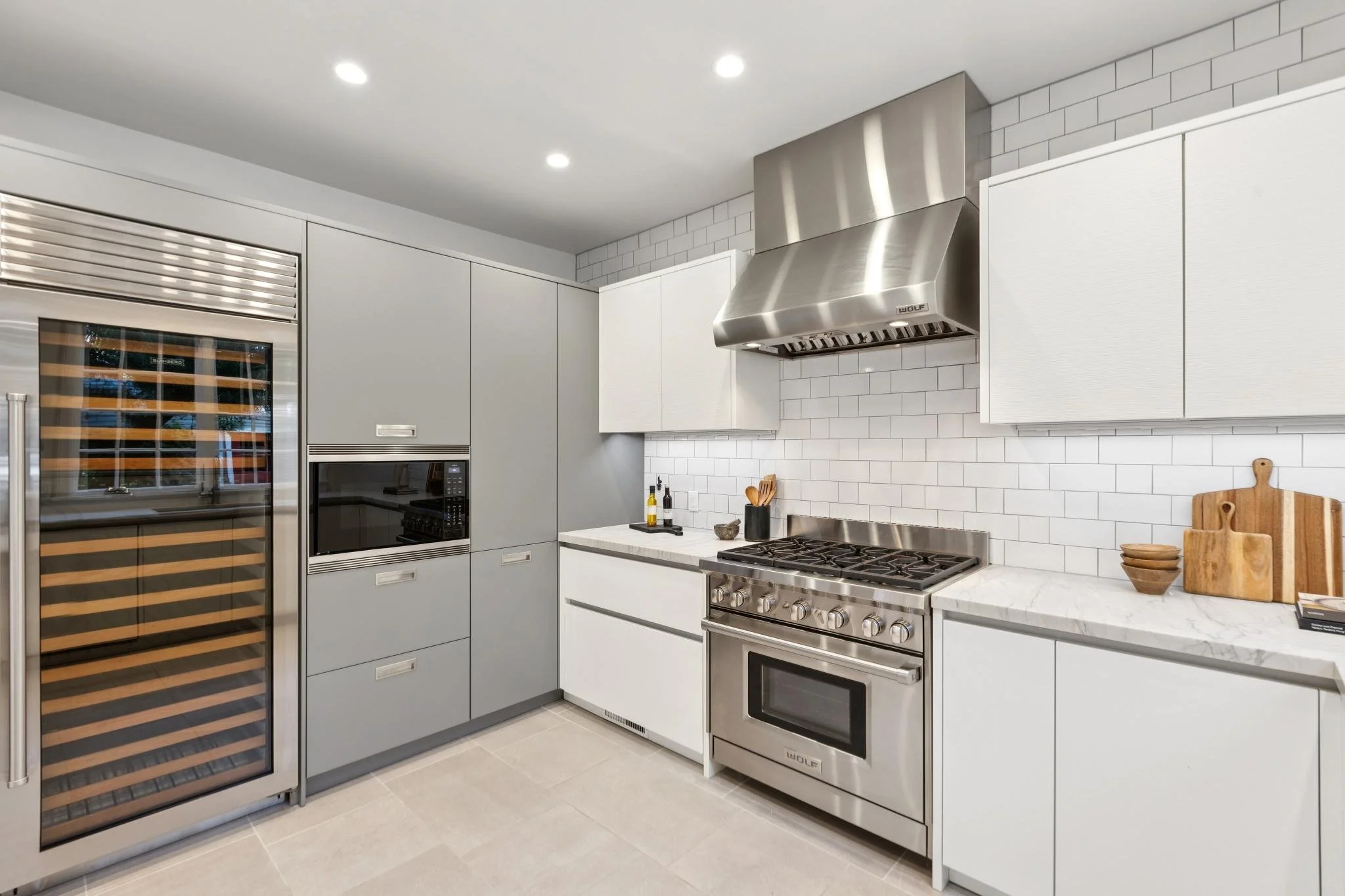 Modern kitchen with gray cabinetry, a stainless steel stove with a range hood, white marble countertops, and a white subway tile backsplash. There is a wine fridge on the left, along with small kitchen accessories and wood cutting boards on the count