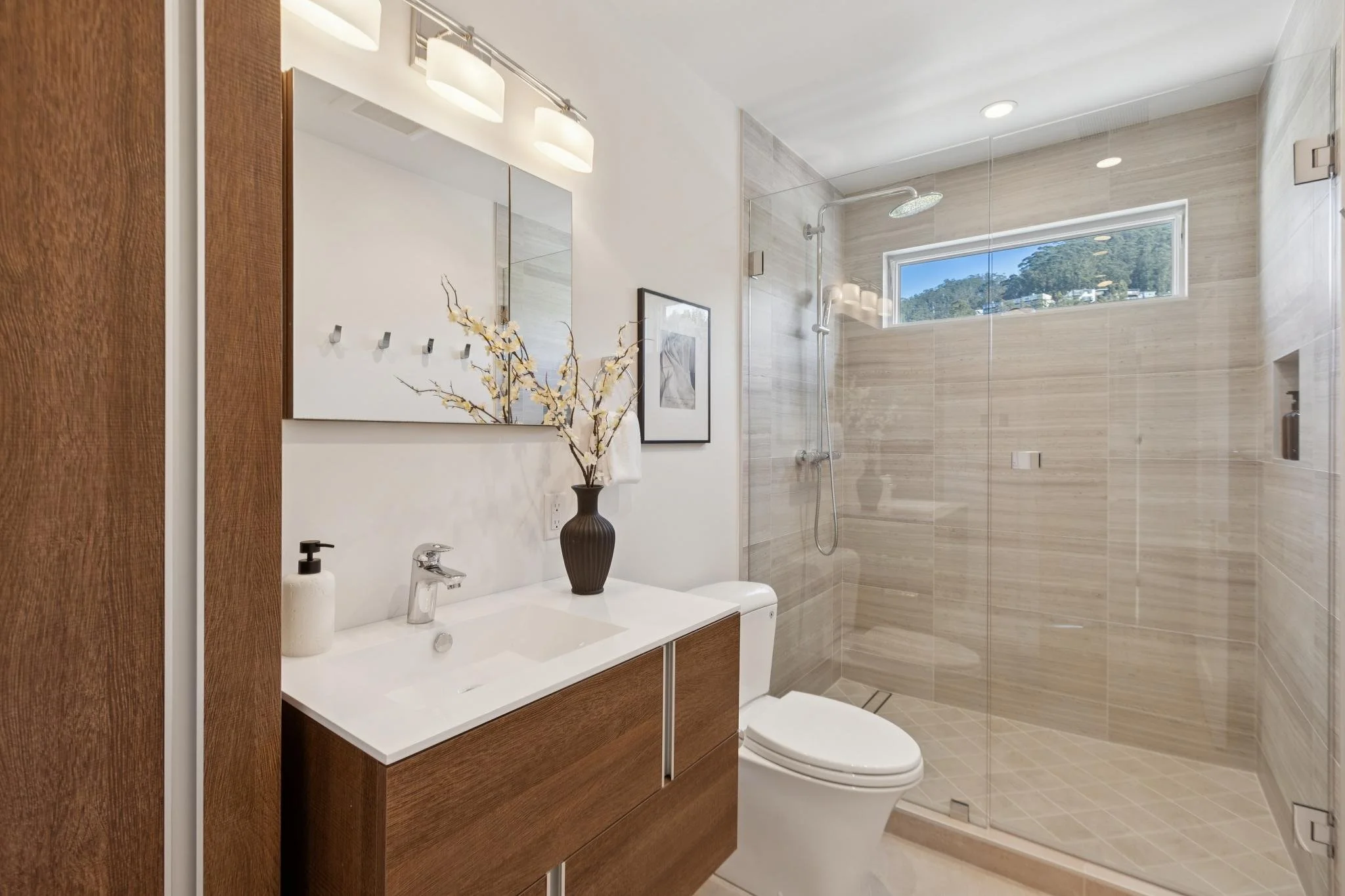 Modern bathroom featuring a wooden vanity with a white countertop, a faucet, and a soap dispenser. A black vase with decorative branches sits on the vanity. The shower area has beige tiles, a showerhead, a built-in niche for toiletries, and a window 