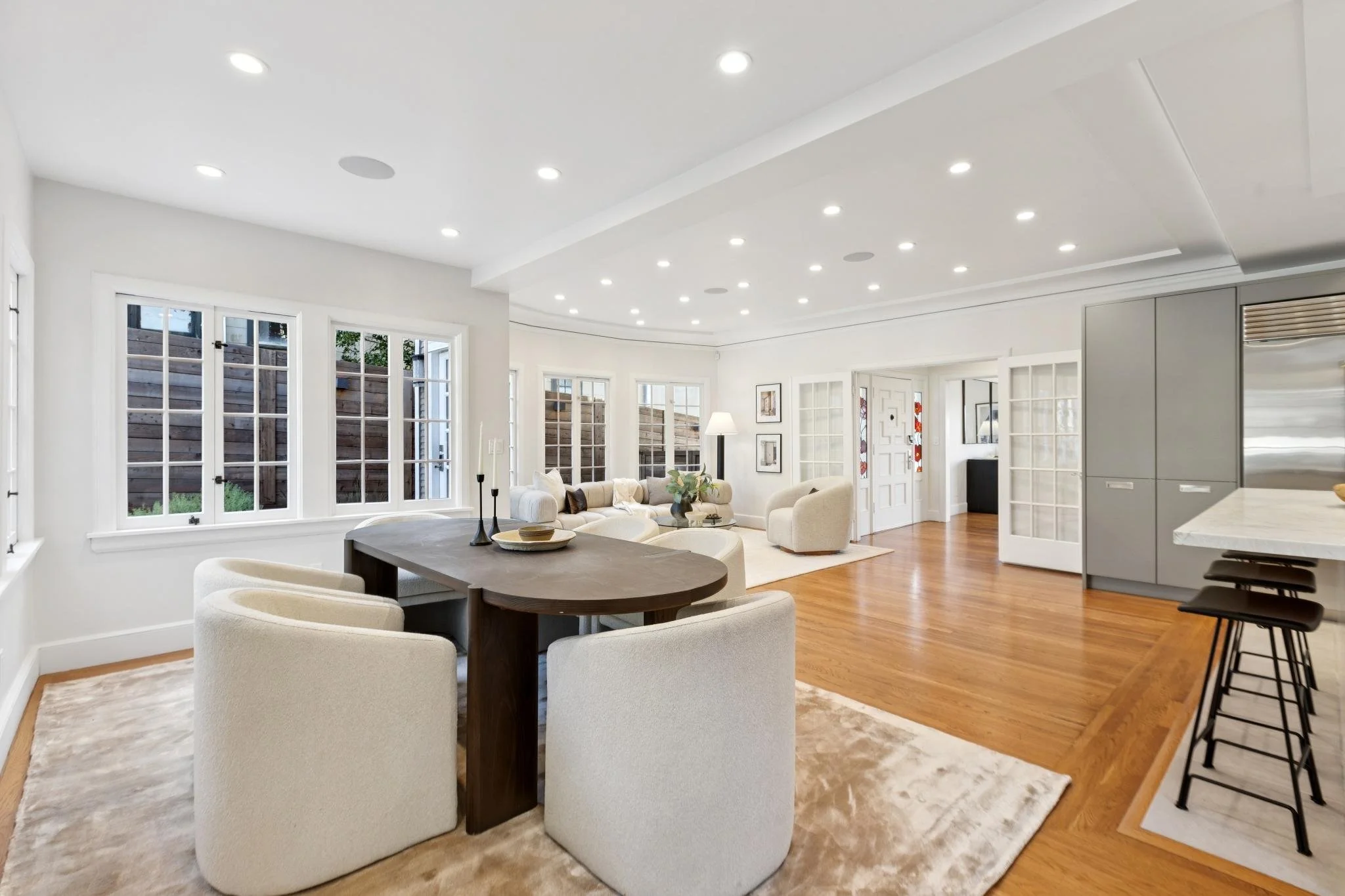Open-concept living and dining area with white walls, hardwood floors, large windows, and recessed lighting. Contemporary furniture includes a white sofa, armchairs, a black dining table with white chairs, and a kitchen with gray cabinets and bar sto
