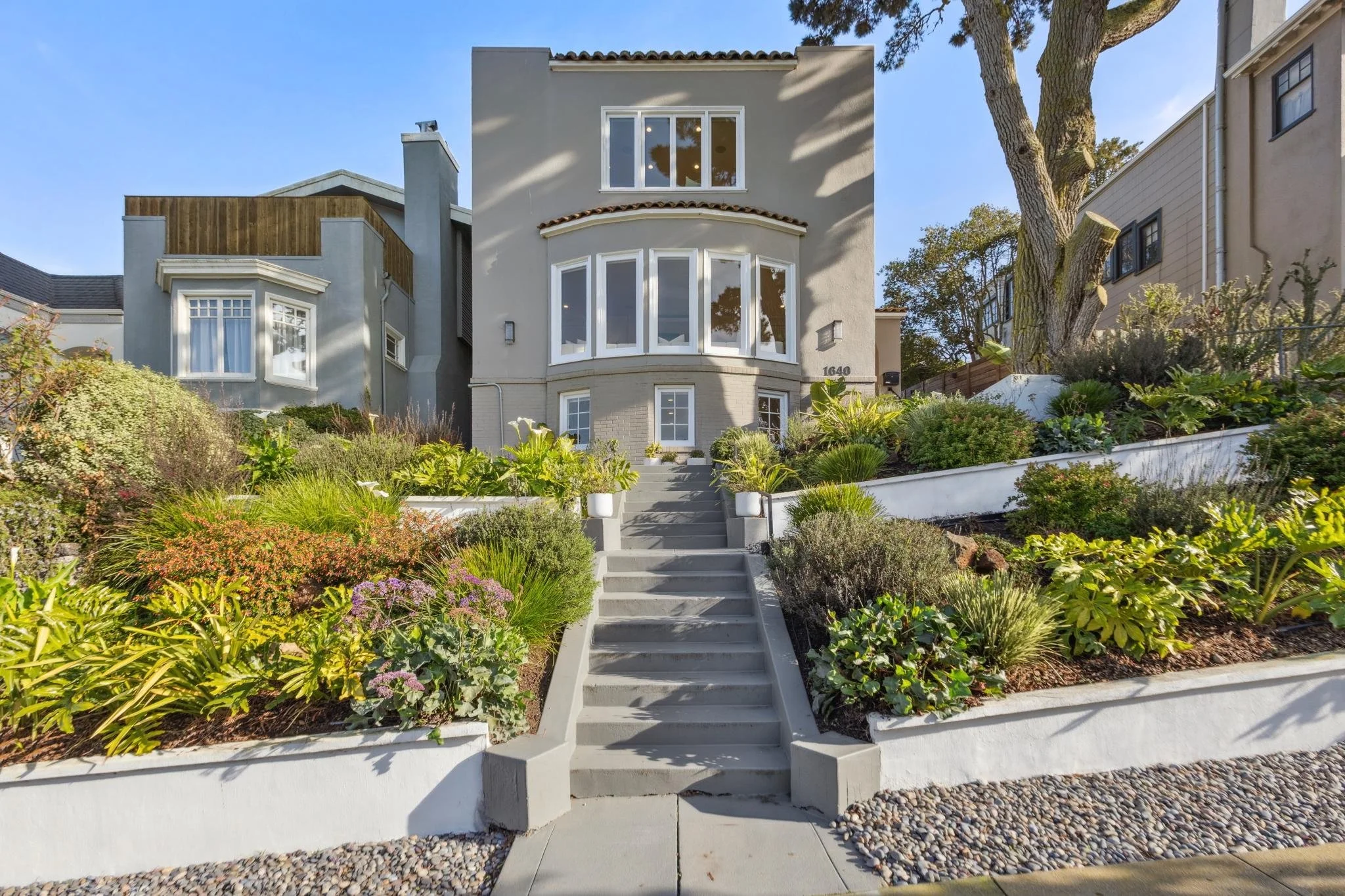A modern multi-story house with a front garden and a staircase leading to the entrance, featuring lush greenery and trees around.
