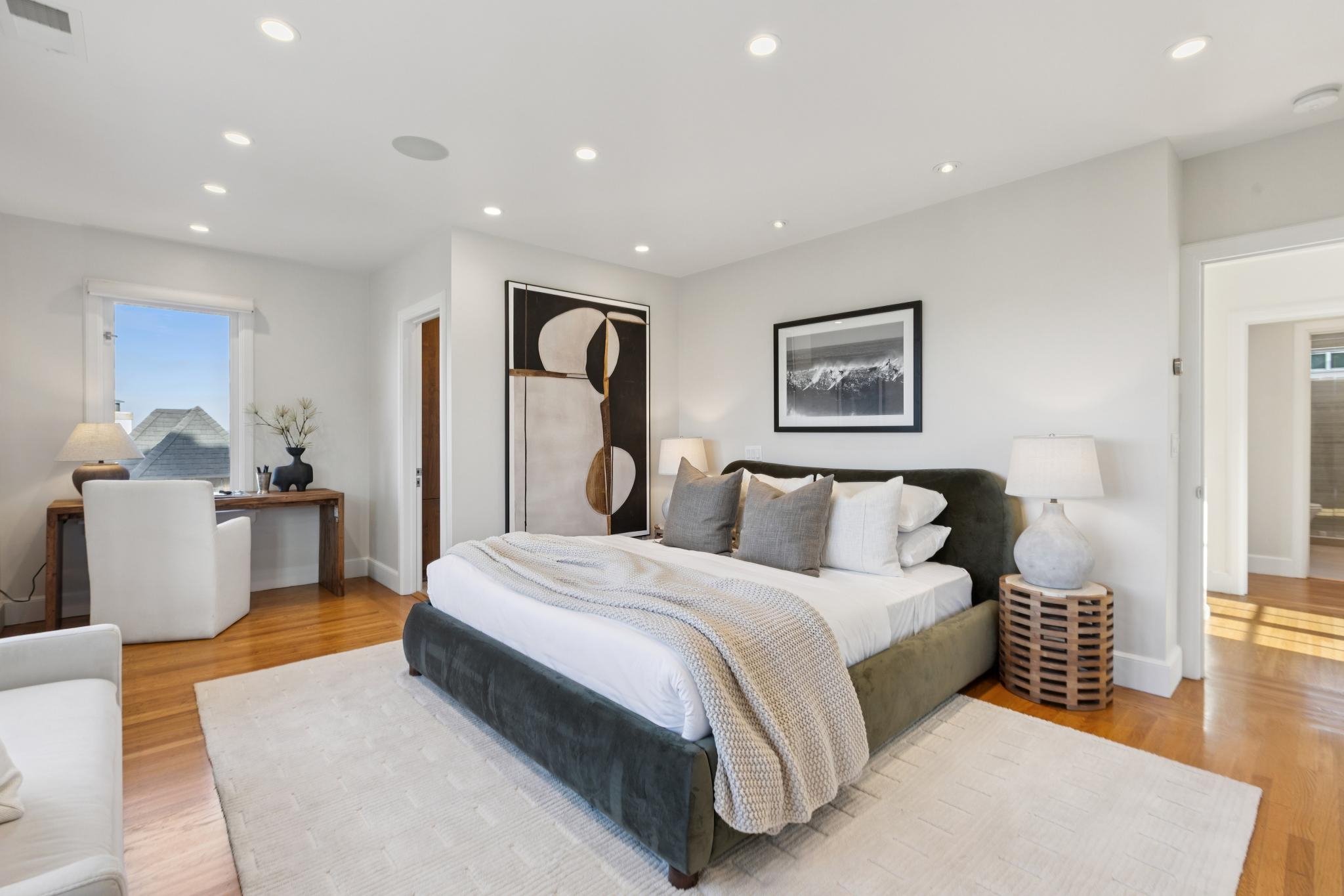 Bright bedroom with white walls, hardwood floor, and modern furniture. Features a large bed with a dark green upholstered frame, white bedding, and multiple pillows. There are two framed artworks above the bed, one with abstract black and white design and the other with a landscape.