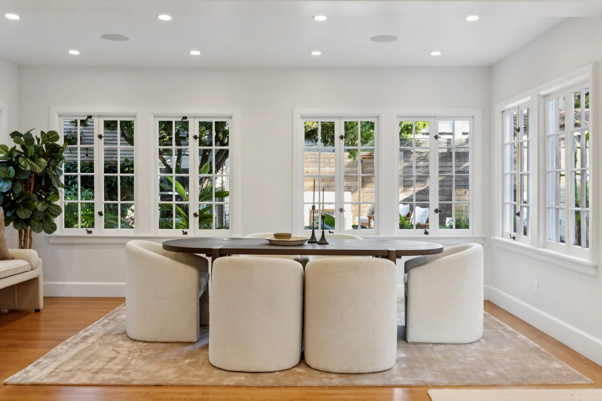 Bright dining room with large windows, dark wood table, six cream upholstered chairs, decorative candlesticks, and greenery outside.