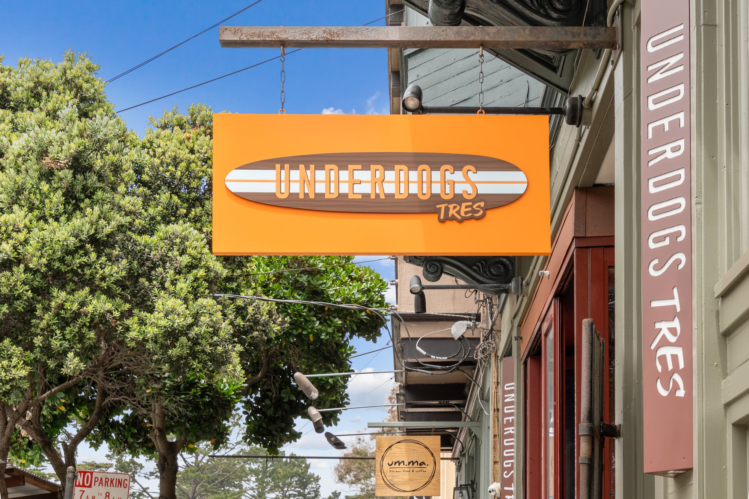 Storefront sign for 'Underdogs Tres' with an orange background and surfboard design, hanging above entrance on a street.