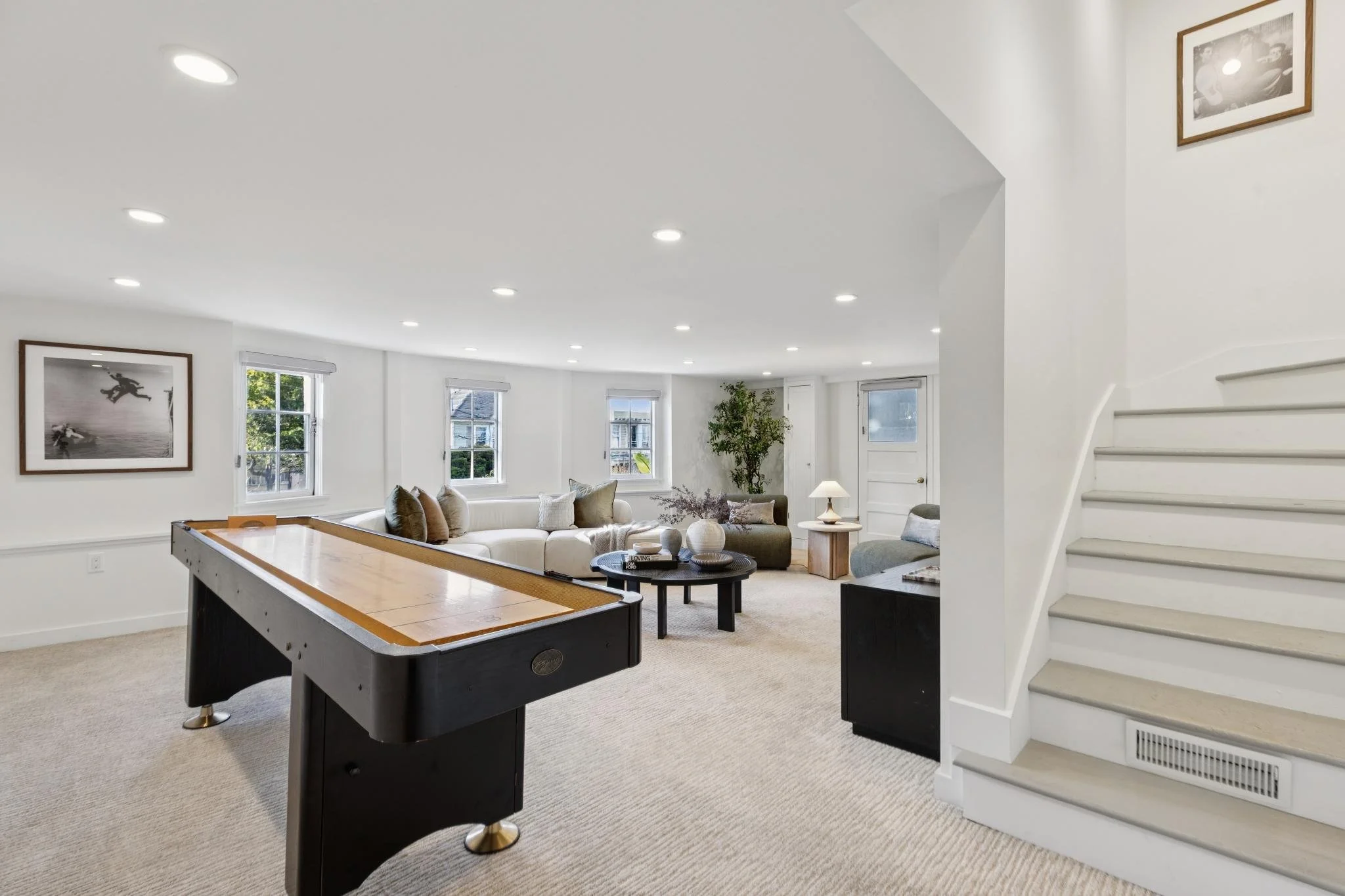 A spacious living room with white walls, carpeted floor, multiple windows, a white sectional couch with pillows, and a dark green armchair. There is a round coffee table with vases and a small side table with a lamp. A staircase with a white banister