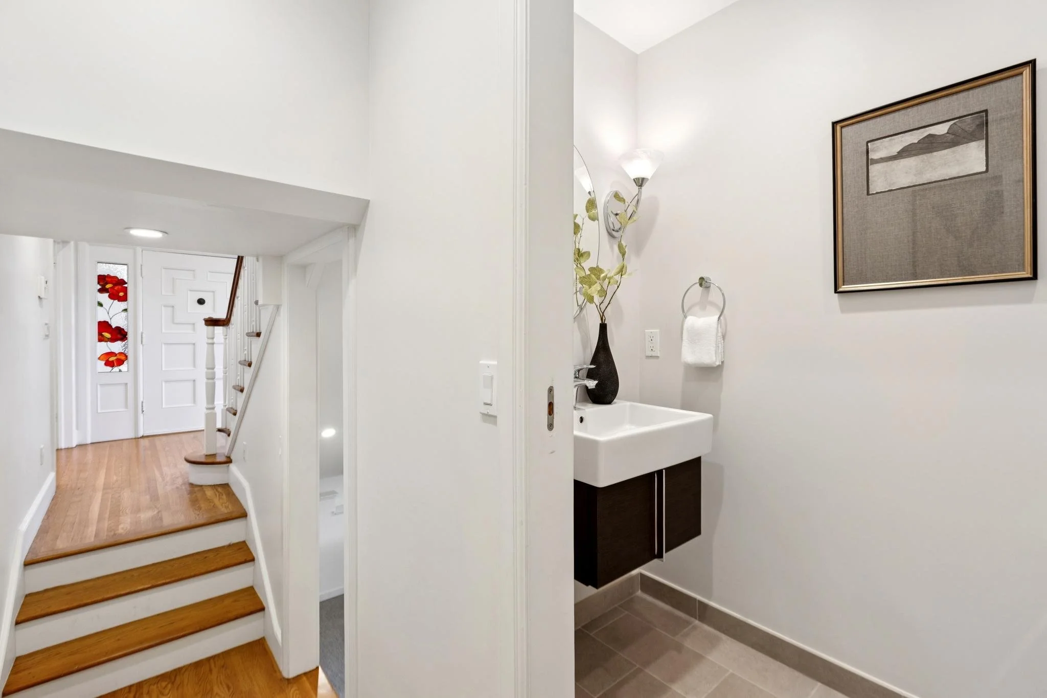 A small bathroom with a white sink, dark cabinet, a towel ring with a white towel, a framed picture on the wall, a tall vase with decorative leaves, and a light fixture. The bathroom is adjacent to a staircase leading to the first floor, with a hallw