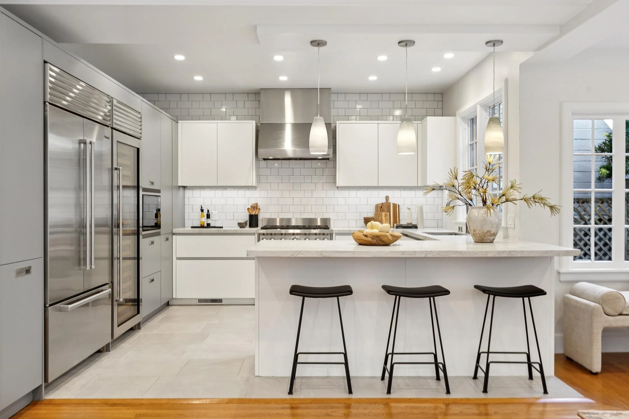Modern kitchen with white cabinets, stainless steel appliances, white subway tile backsplash, and a white island with black barstools. Decor includes a vase with dried branches and a bowl of fruit.