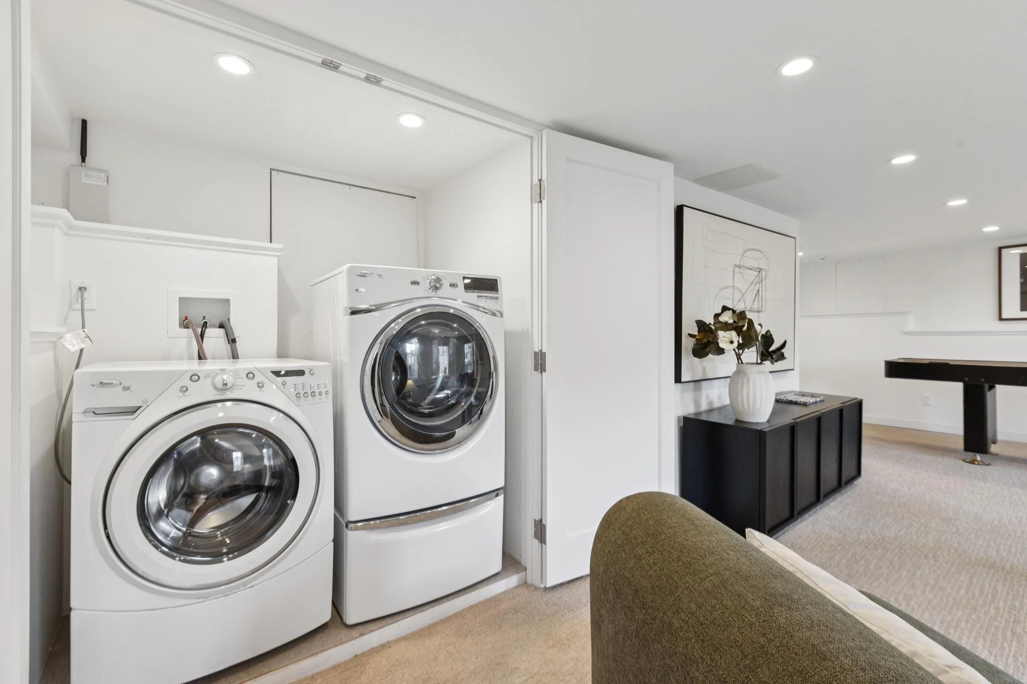 Laundry area with white washer and dryer in a small closet, adjacent to a living space with decorated black cabinet, framed artwork, flower vase, and ceiling lights.