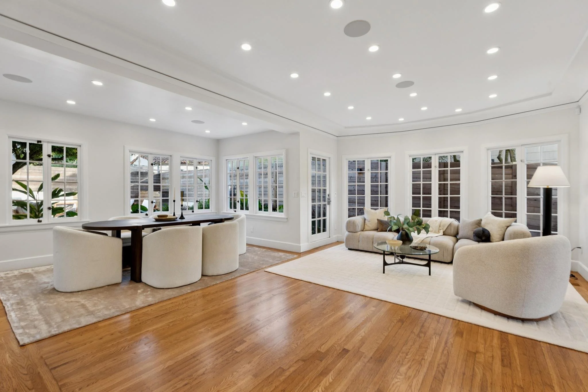 Bright living room with large white sectional sofa, glass coffee table, and floor lamp in front of multiple windows showing an outdoor wooden fence. Adjacent dining area with dark wood table and cream upholstered chairs, situated on beige rugs over h