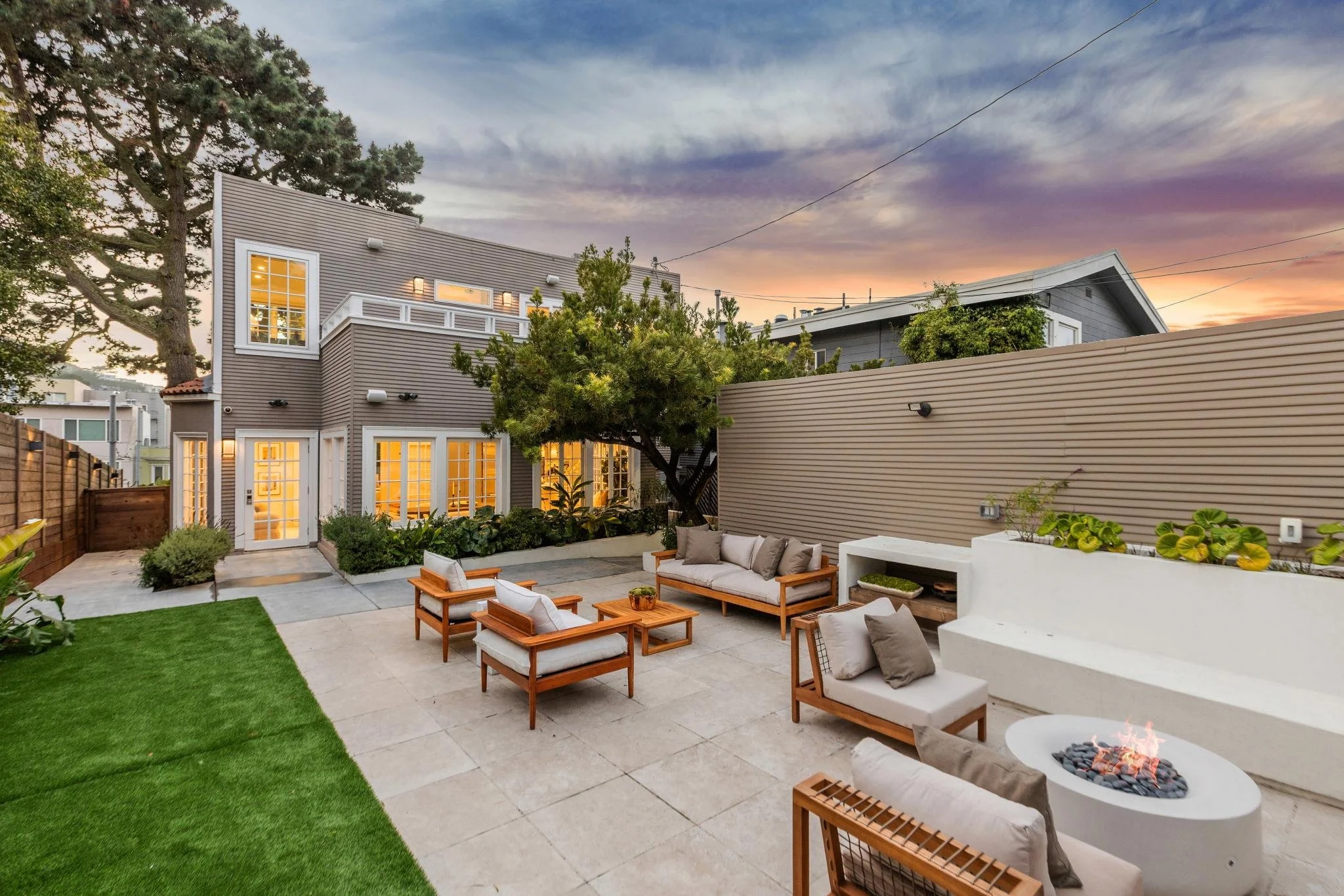Outdoor patio with seating area, fire pit, and garden at dusk, with a modern house in the background.