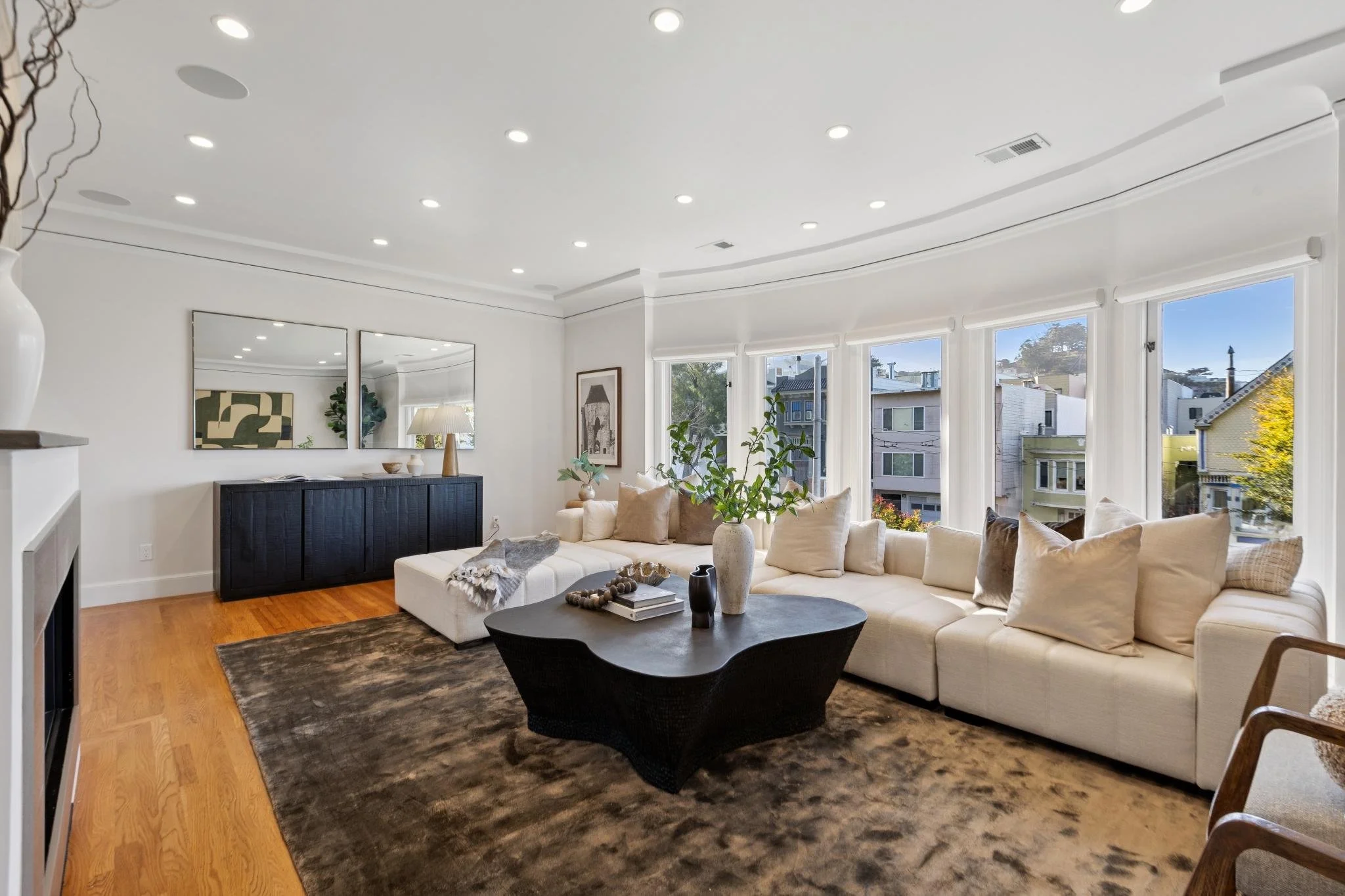 Bright living room with large bay windows, white sectional sofa with beige and brown cushions, black coffee table with decorative vases and books, brown area rug, and black sideboard with mirrors, inside a modern home.