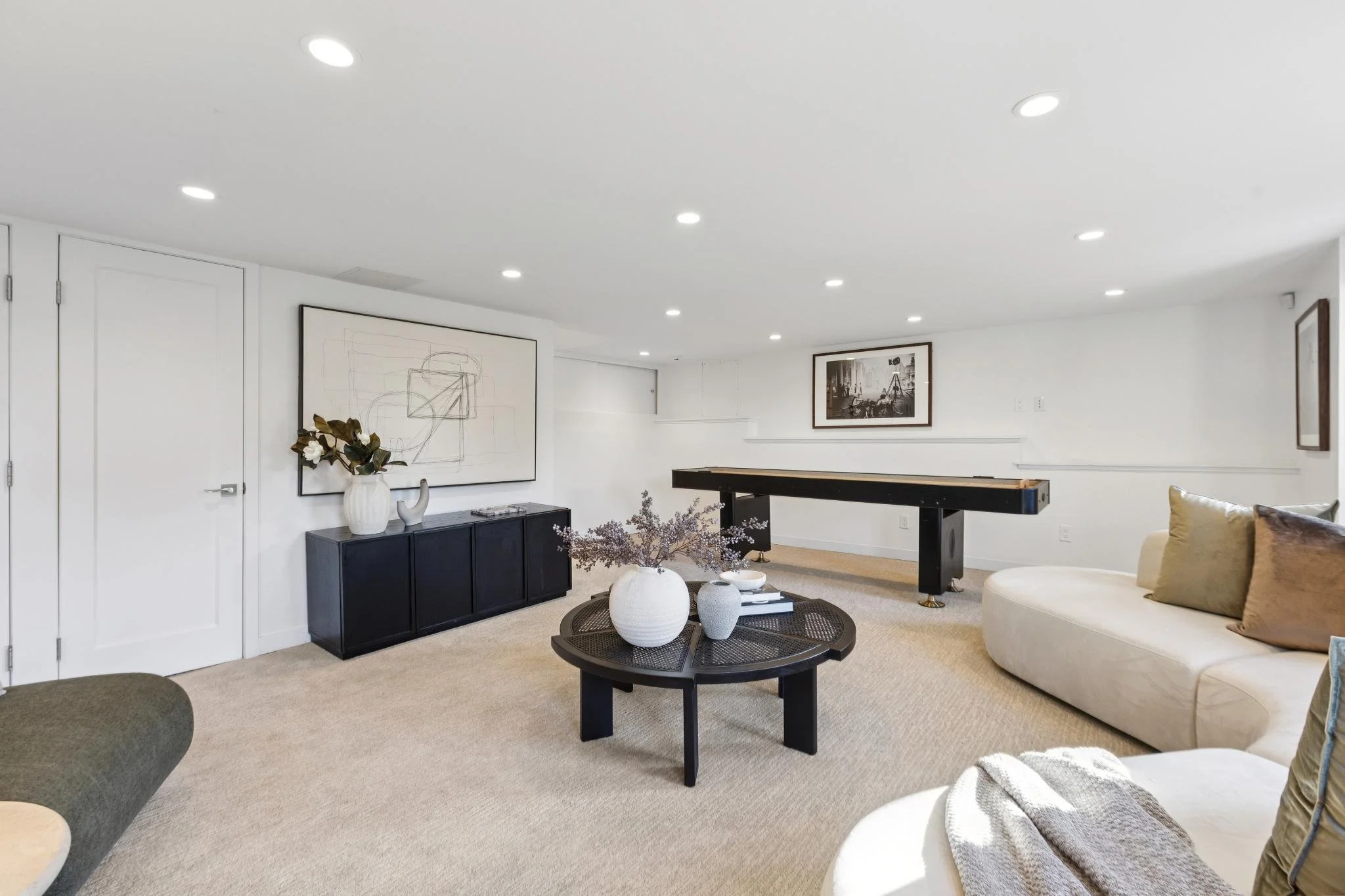 A spacious, well-lit living room with white walls, beige carpet, black and white artwork, and a pool table. It has modern furniture including a curved cream sofa with pillows, a black coffee table with decorative vases, and a black sideboard with a l