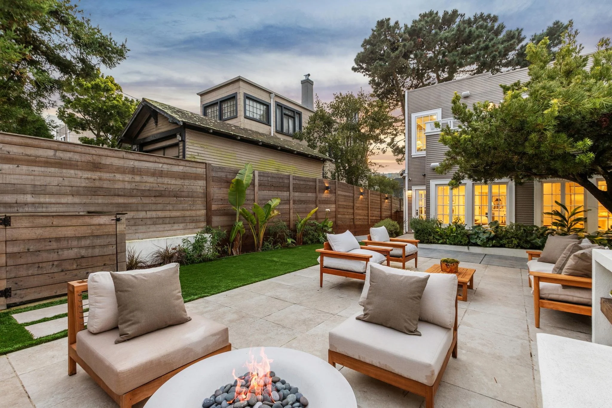 A backyard patio at dusk with outdoor seating including armchairs and a fire pit, surrounded by a wooden fence, greenery, and a multi-story house with lit windows.