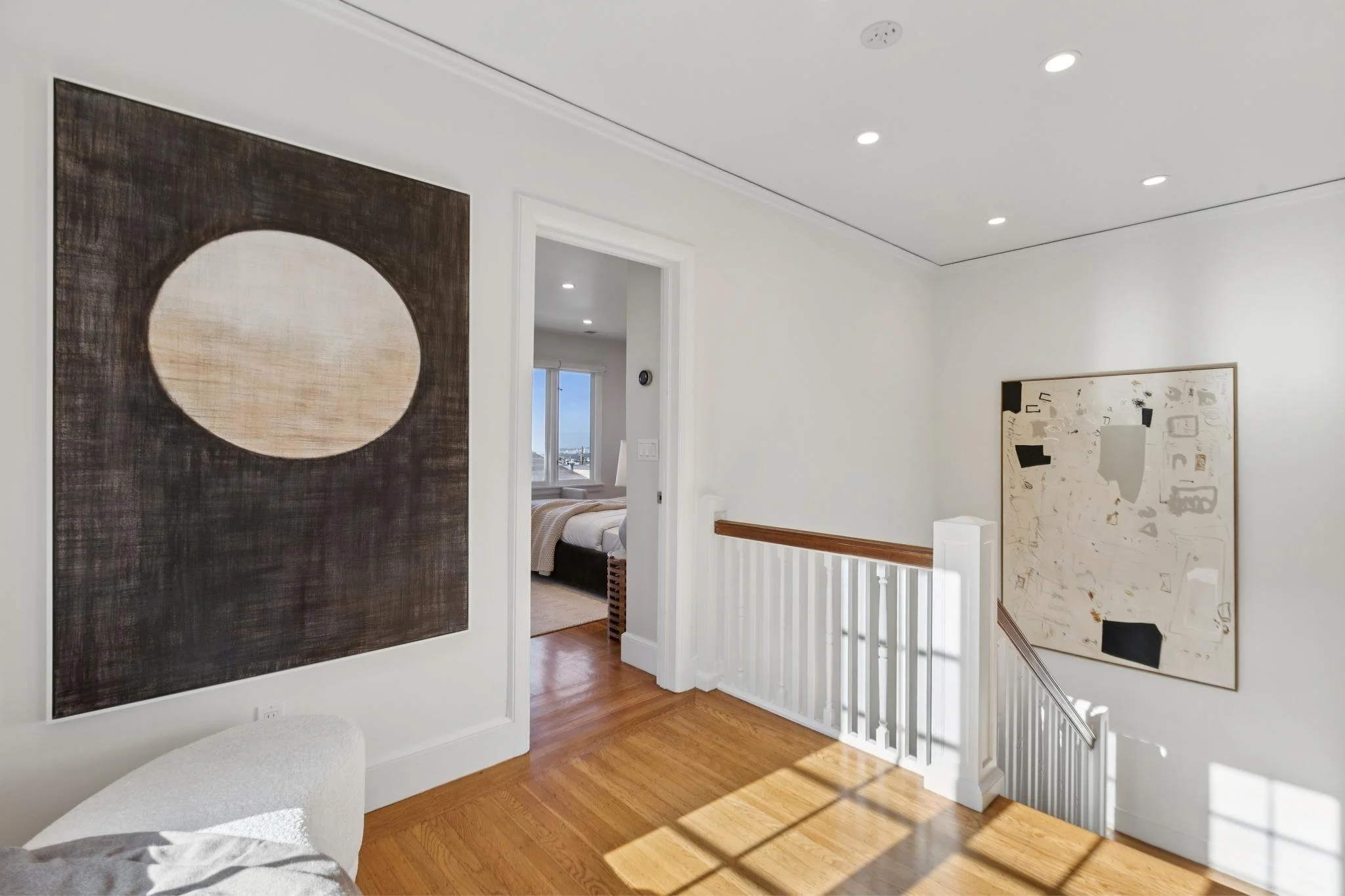 Bright hallway with hardwood floors, abstract art on white walls, and a view into a room with a bed and large window.