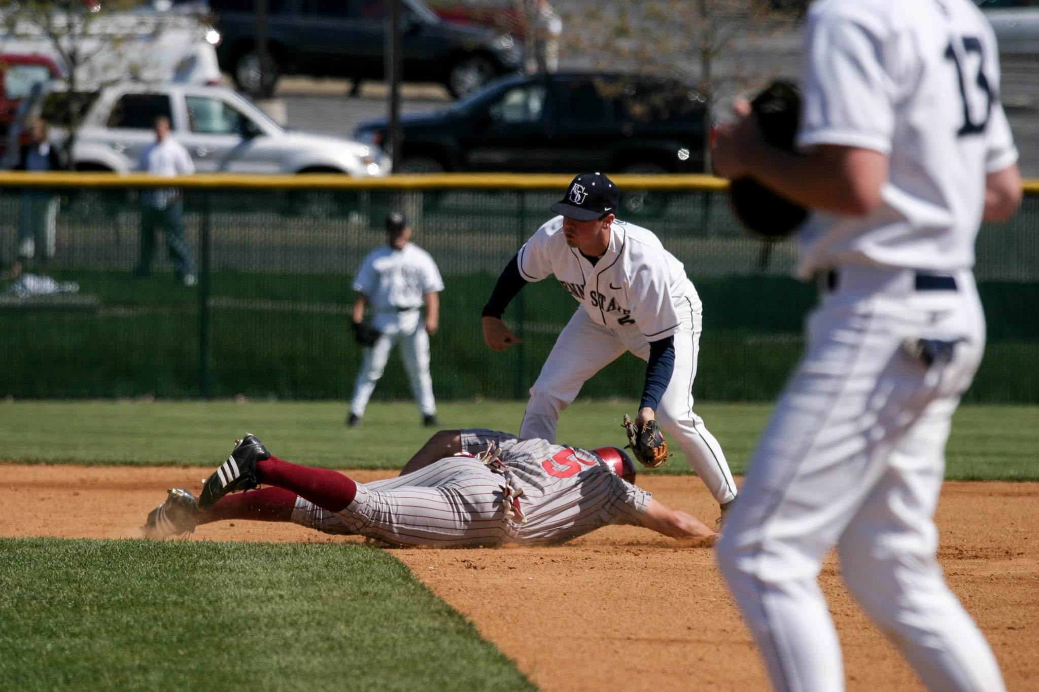 A baseball player is sliding into a base while another player attempts to tag him out during a game on a baseball field, with a coach or teammate nearby.