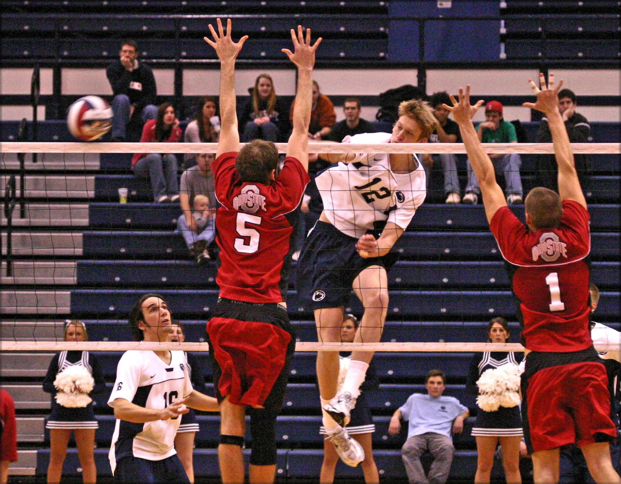 A volleyball game with a player in a white jersey spiking the ball over the net while two players in red jerseys attempt to block it. Spectators sit in the background in a gymnasium.