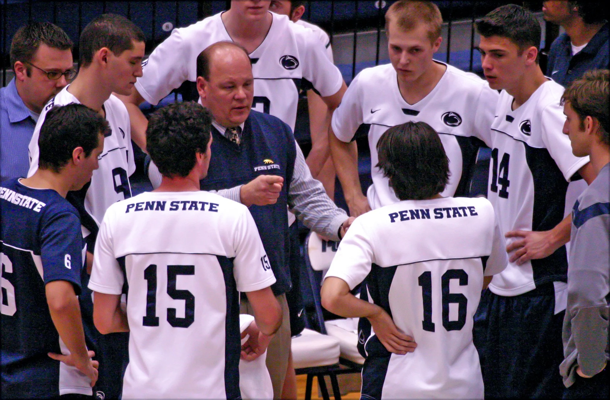 Penn State men's volleyball team in a huddle listening to their coach during a game or practice, with players wearing white and navy uniforms with the Penn State logo and numbers.