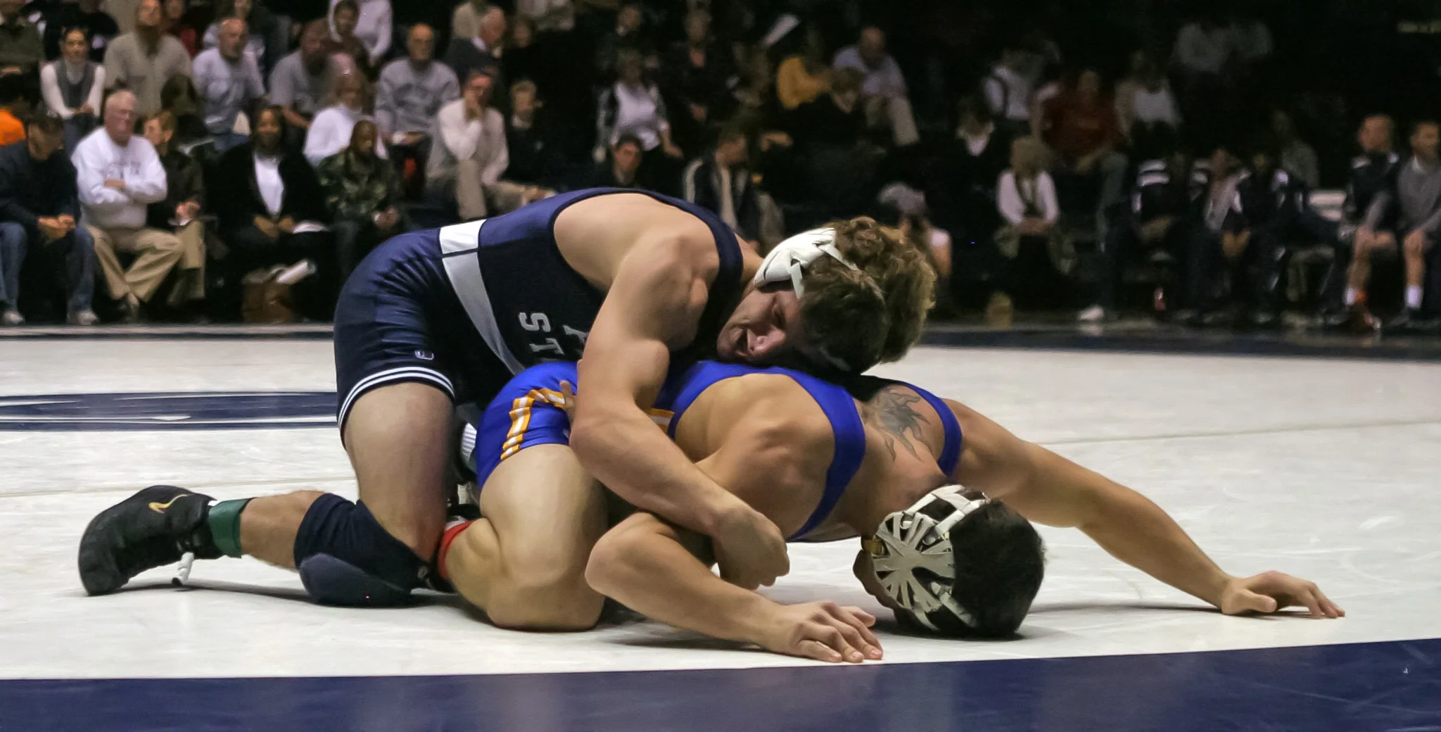 Two wrestlers competing on a wrestling mat, one in a blue singlet and the other in a black singlet, with a crowd of spectators watching in the background.