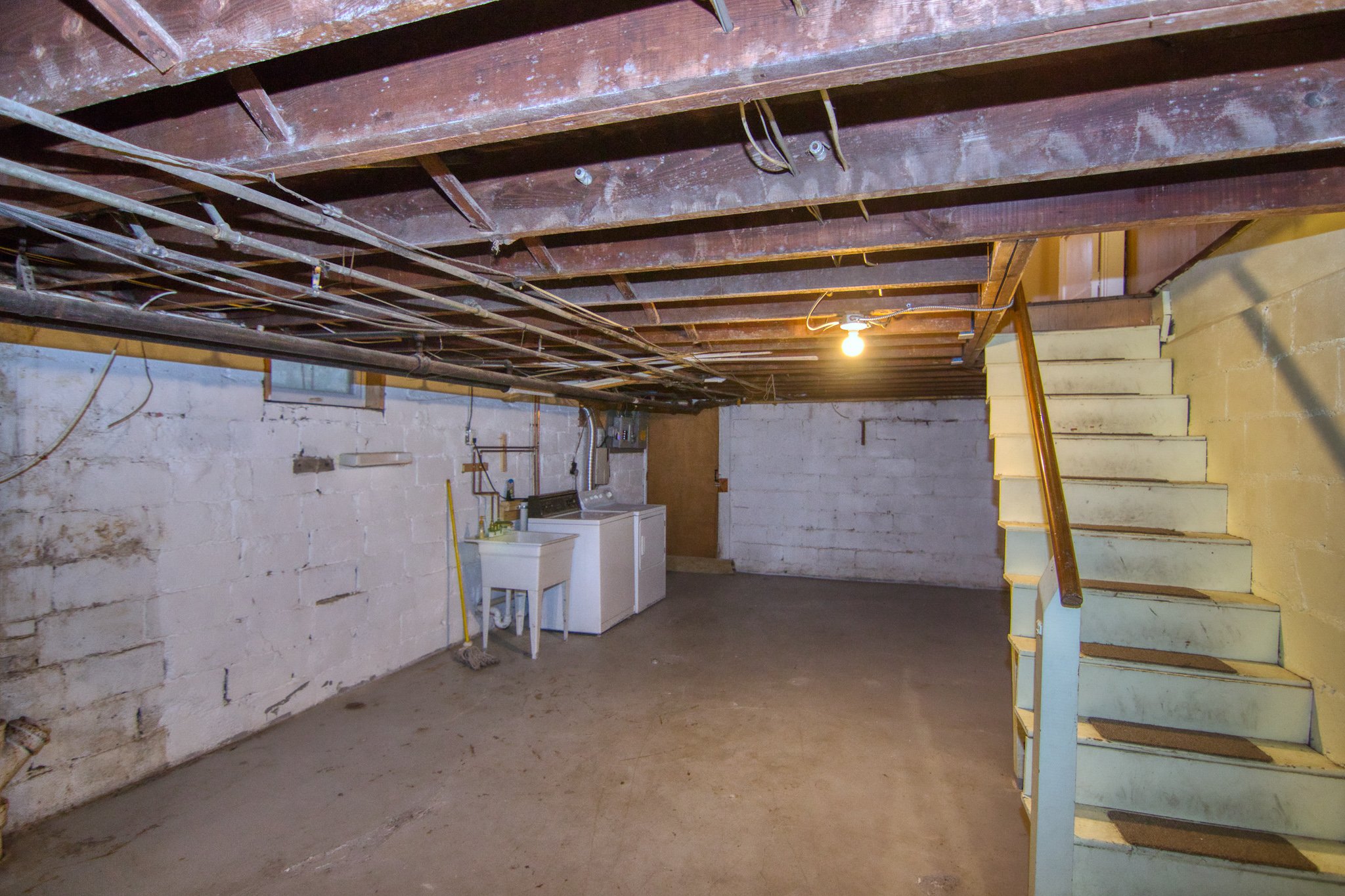 Basement with exposed ceiling beams, washing machine and utility sink, white brick walls, concrete floor, and stairs leading up to the right.