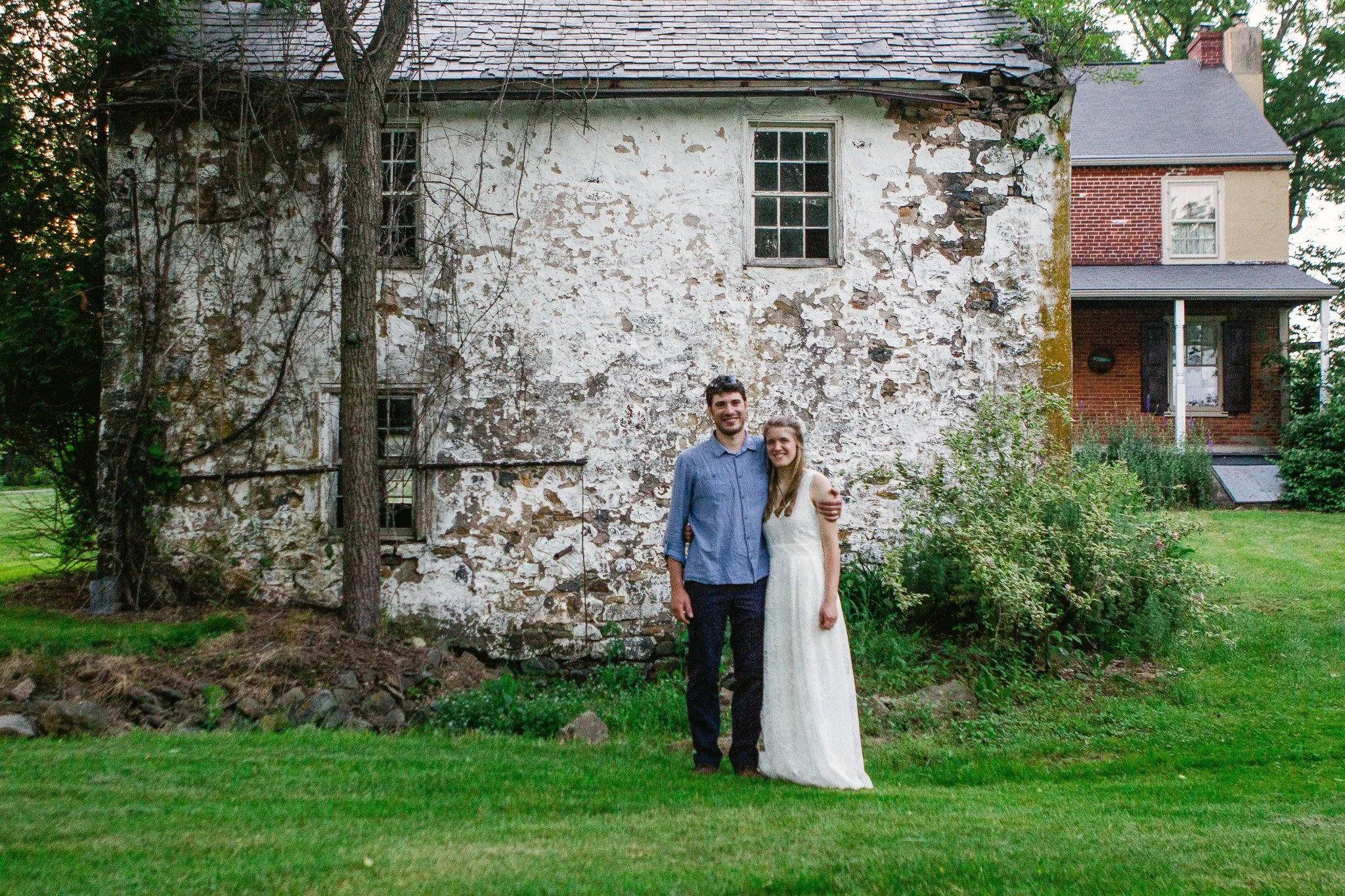 A young couple, with the man in a blue shirt and the woman in a white dress, standing close together with their arms around each other in front of a worn, white stone house with peeling paint. Green grass and some trees surround them.