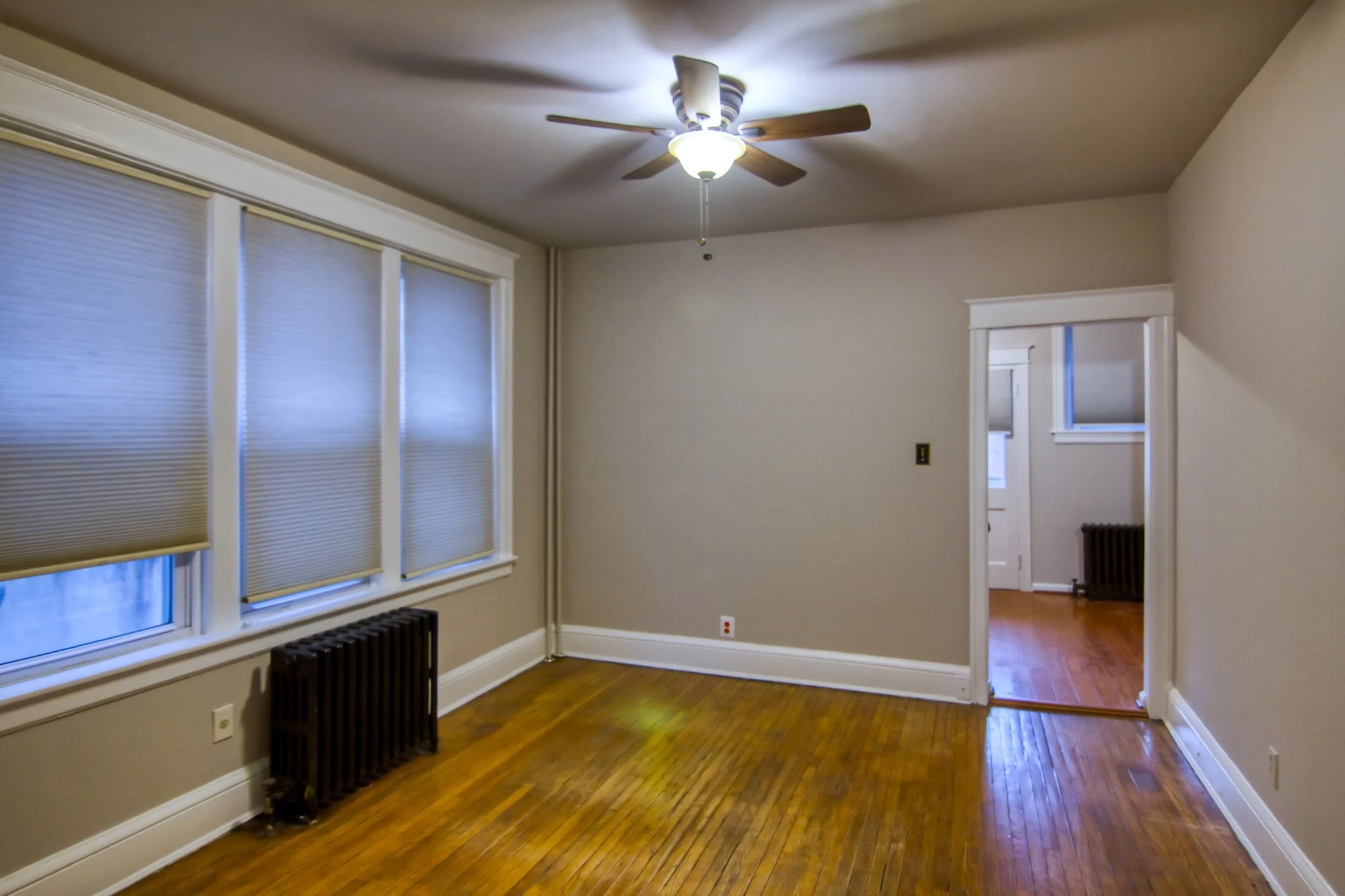 Empty room with hardwood floors, beige walls, large windows with blinds, white crown molding, a ceiling fan, and a doorway leading to another room.