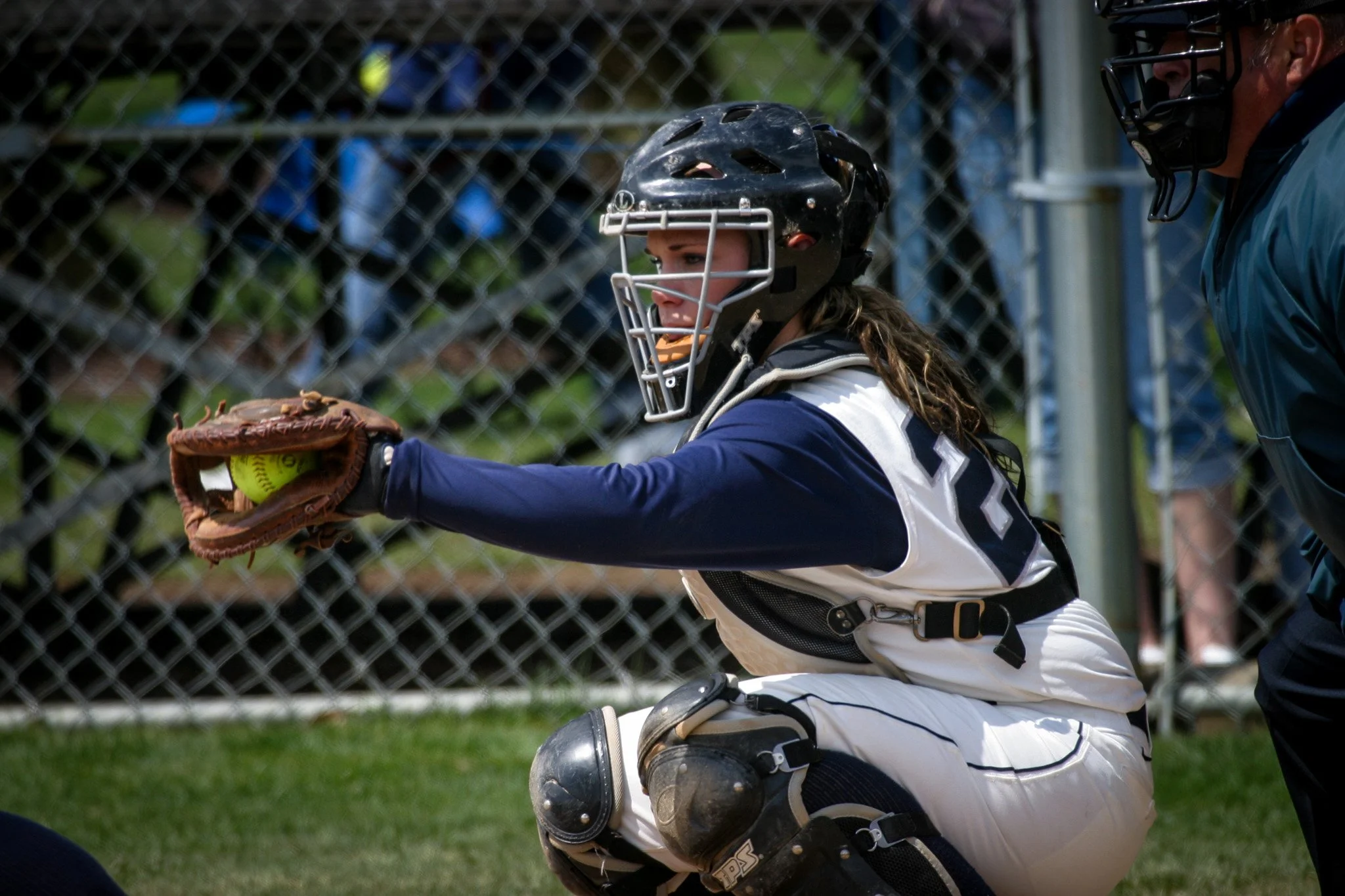A female softball catcher in gear reaching out to catch a green softball with a brown glove during a game.