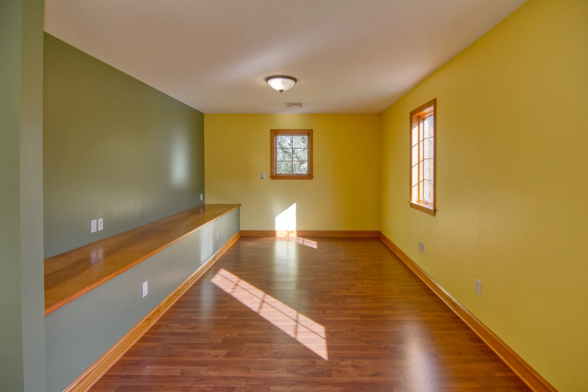 Empty room with hardwood floors, light green and yellow walls, three windows with wooden frames, and a ceiling light fixture.