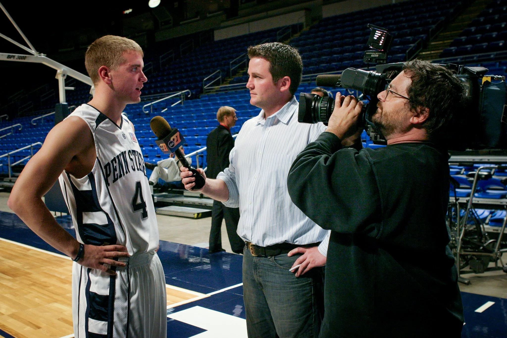 A basketball player from Penn State being interviewed by a reporter, while a cameraman records the interview inside an empty basketball arena.