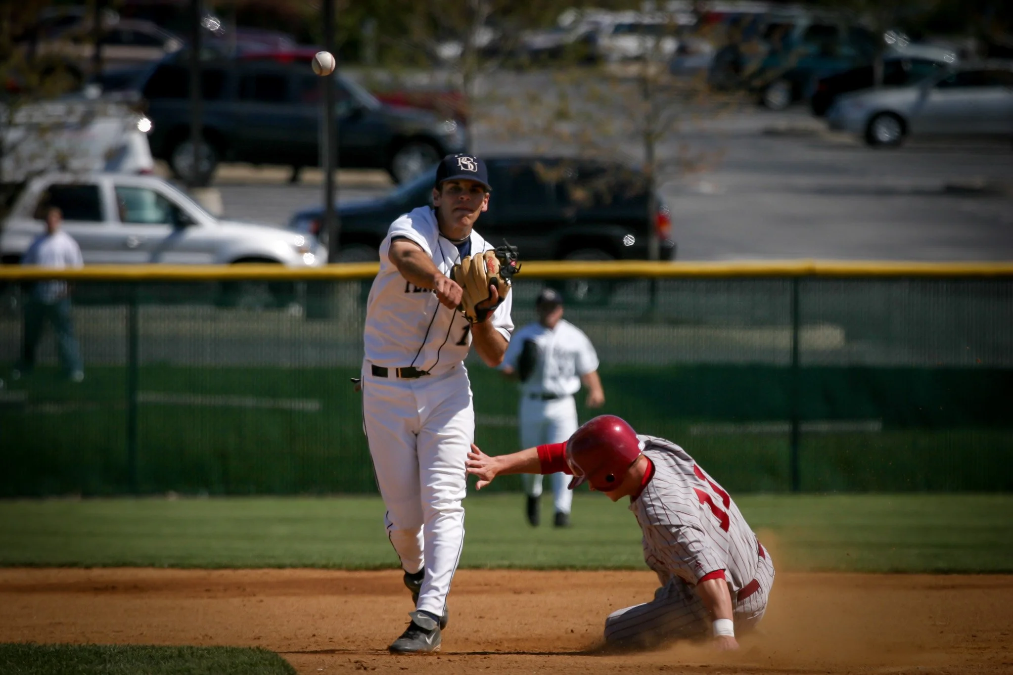 A baseball player in a white uniform is running to second base while a player in a striped uniform with a red helmet is on the ground reaching for the ball during a game.