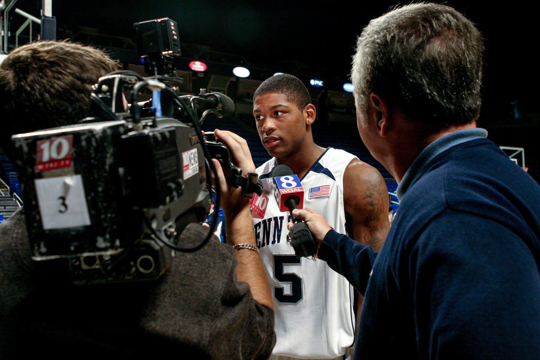 A young male basketball player dressed in a white jersey with the number 5 and a Penn State logo, being interviewed by journalists with microphones in a basketball arena.