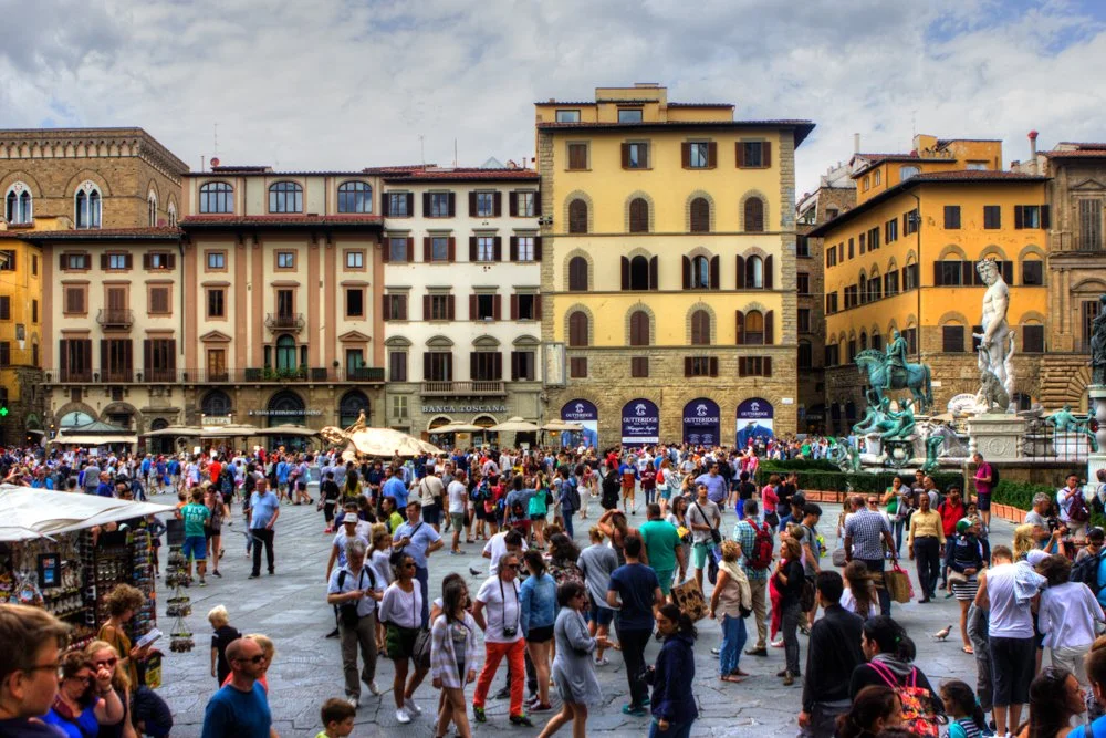 Crowded square in Florence, Italy with historic buildings, statues, and street vendors.