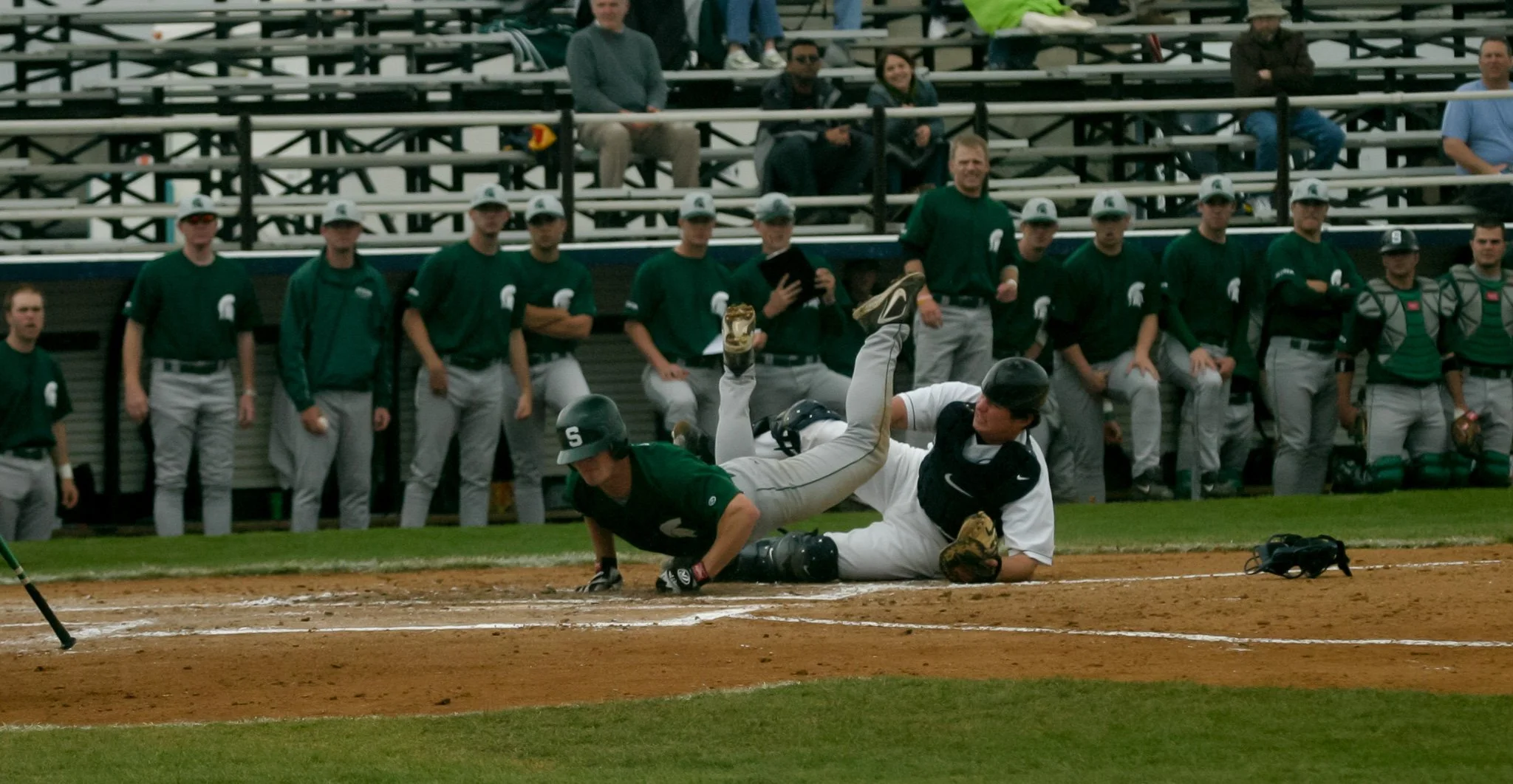 A baseball player sliding into home plate while the catcher attempts to tag him out. Several teammates and spectators are visible in the background.
