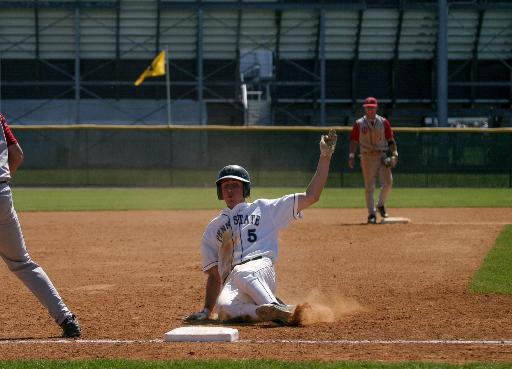 A baseball player from Penn State slides into a base as the baseman reaches to tag him out, on a baseball field during a game.