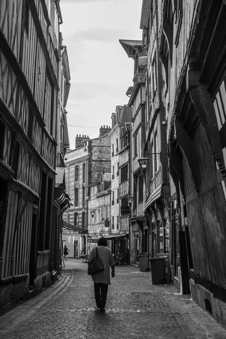 A person walking down a narrow cobblestone street between tall, older buildings with half-timbered and brick facades, possibly in a historic European city.