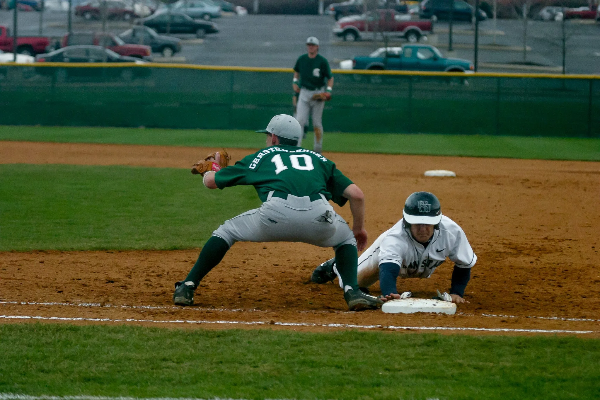 A baseball player in a green jersey and white helmet is tagged out at first base by a player in a white jersey and black helmet. The game is taking place on a baseball field with dirt and grass, and there is an umpire in the background.